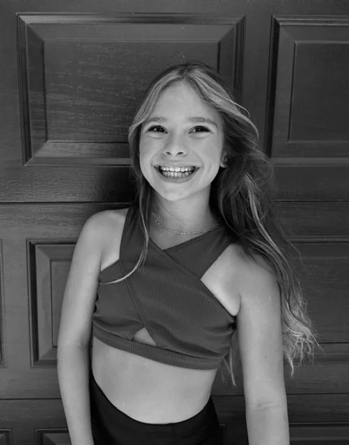 A young girl smiling, standing in front of a wooden door, wearing a sleeveless crop top and a necklace.