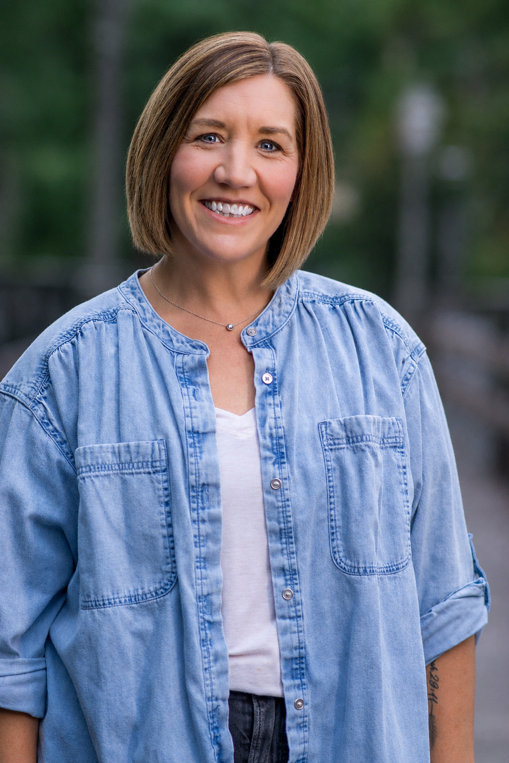 A woman with shoulder-length light brown hair smiling outdoors, wearing a light blue denim shirt over a white T-shirt, with a blurred green background.
