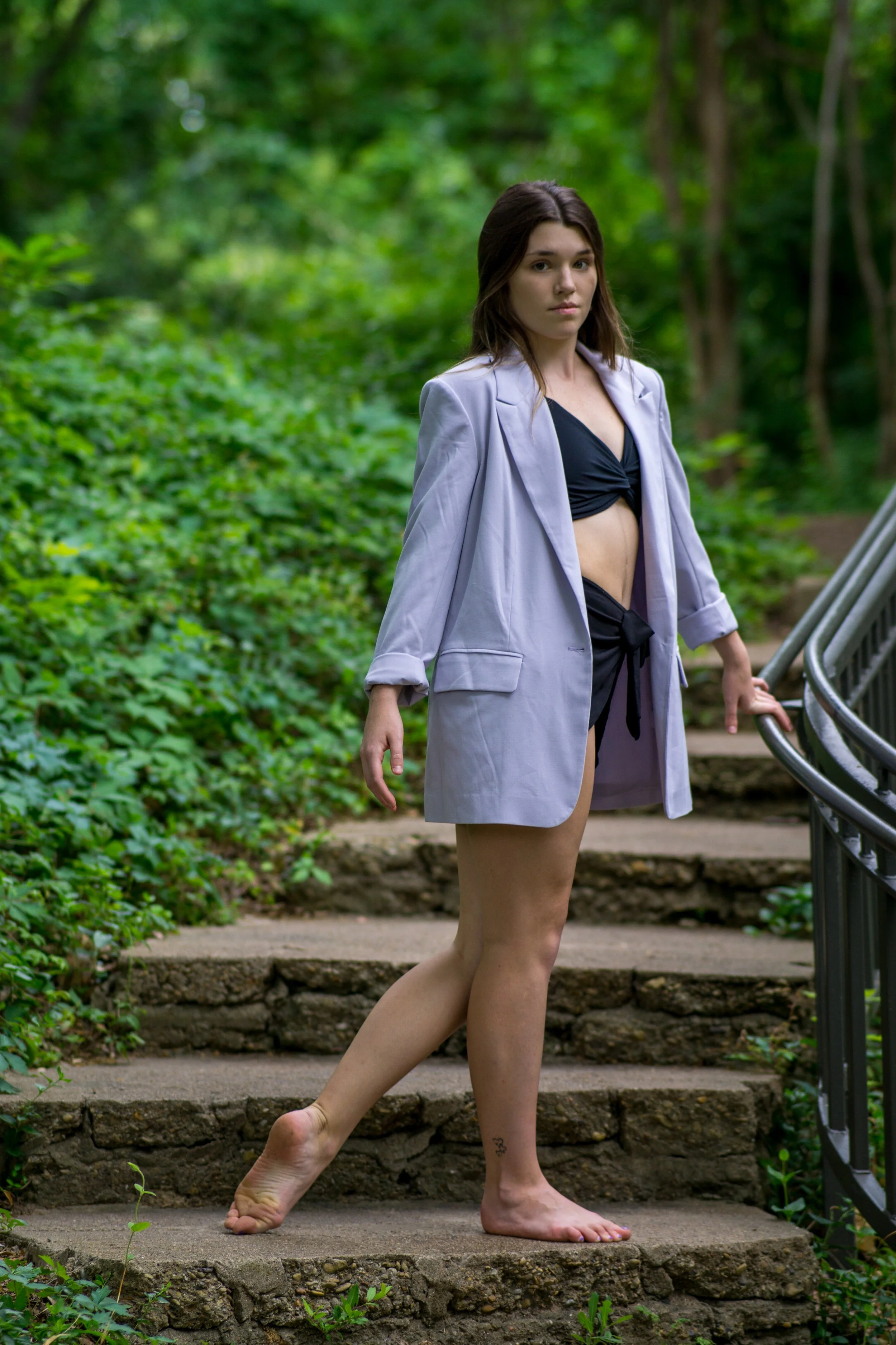 A young woman with brown hair standing on outdoor stone steps surrounded by green foliage, wearing a blazer and black crop top, with one foot on a higher step and one hand holding a railing.