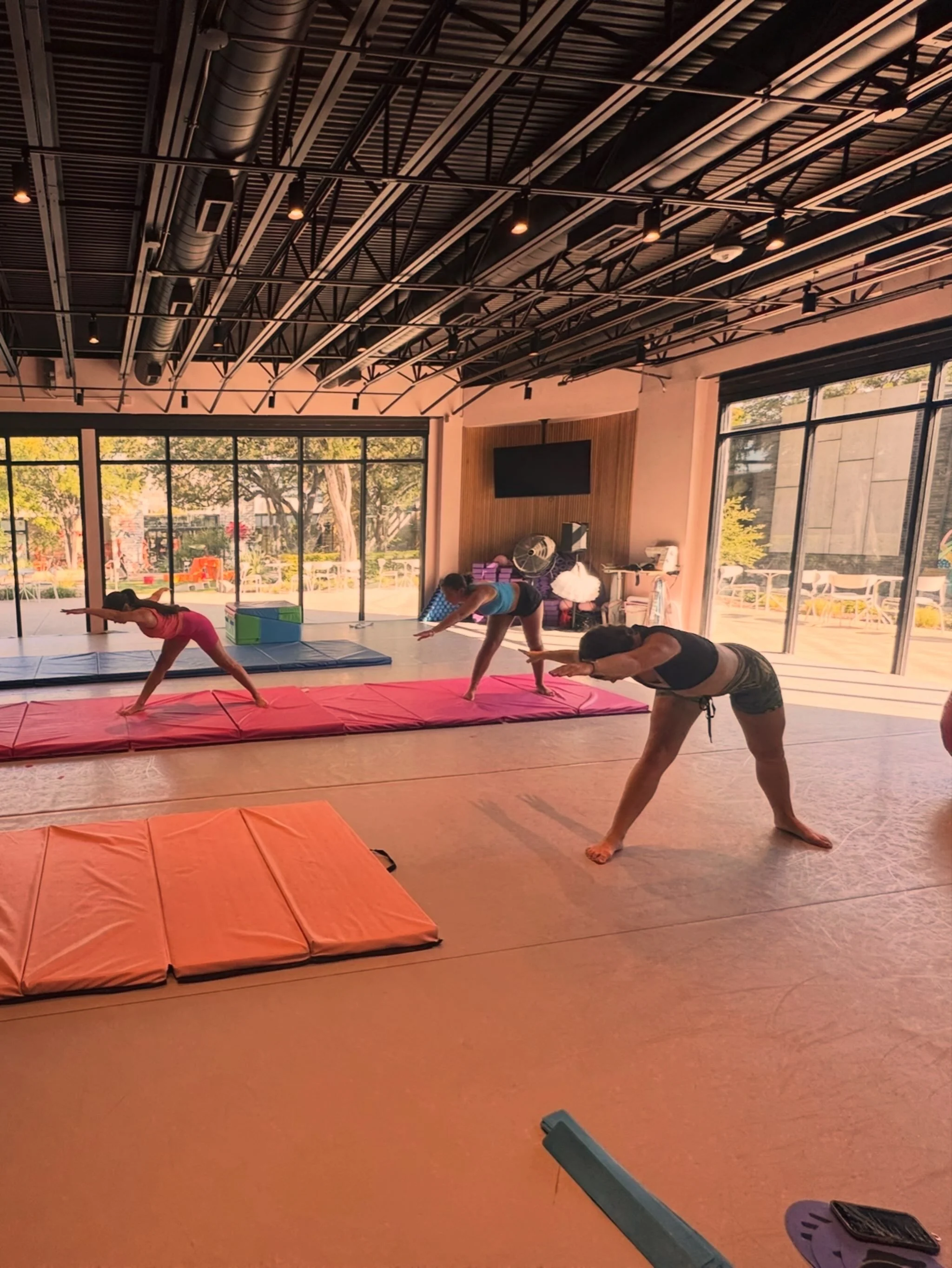 People practicing yoga in a bright, spacious room with large windows showing trees outside. The room has a high ceiling with exposed beams and ductwork. There are yoga mats on the floor, and some people are stretching and balancing.