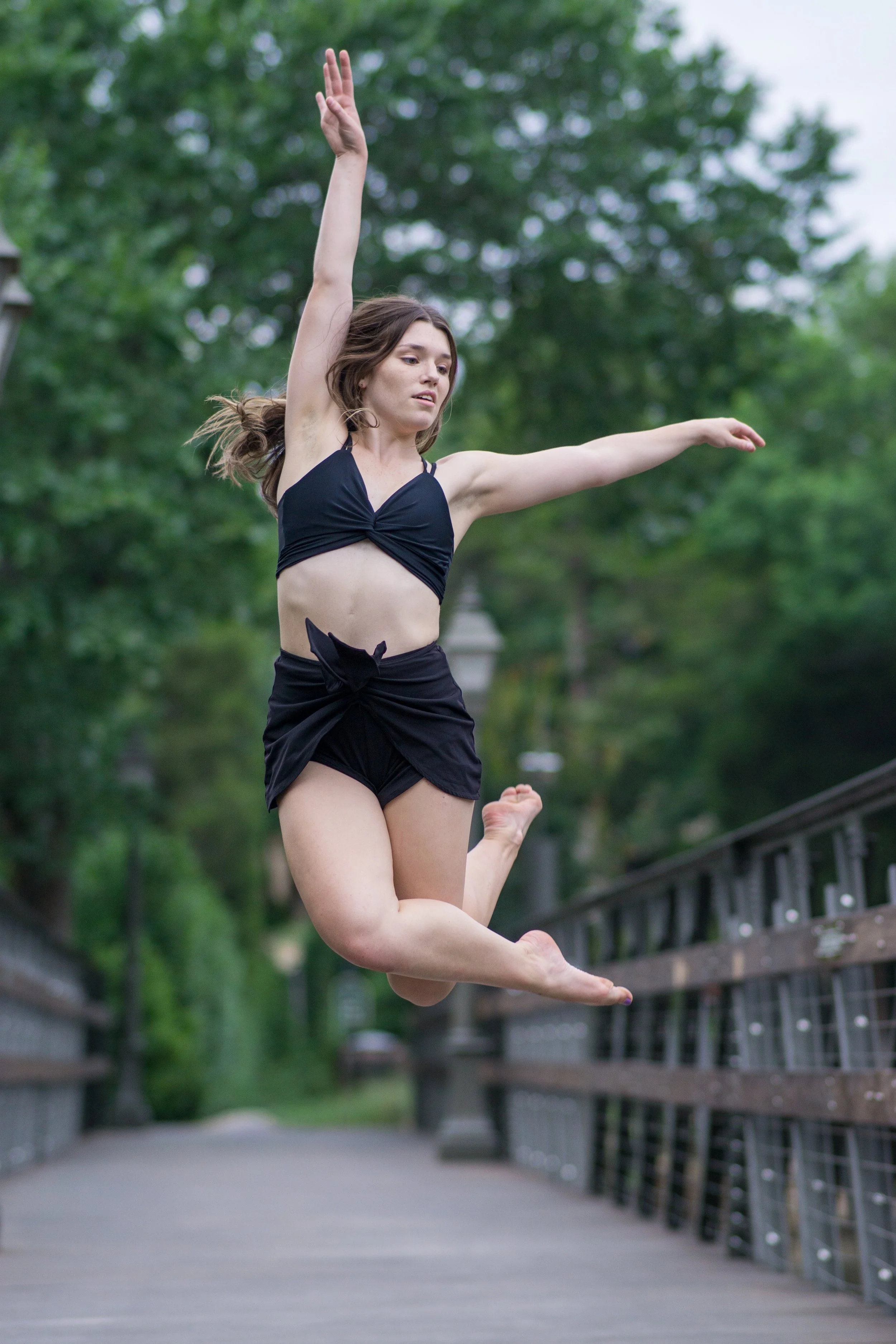 A young woman in black athletic clothing jumping mid-air outdoors on a wooden bridge with green trees in the background.