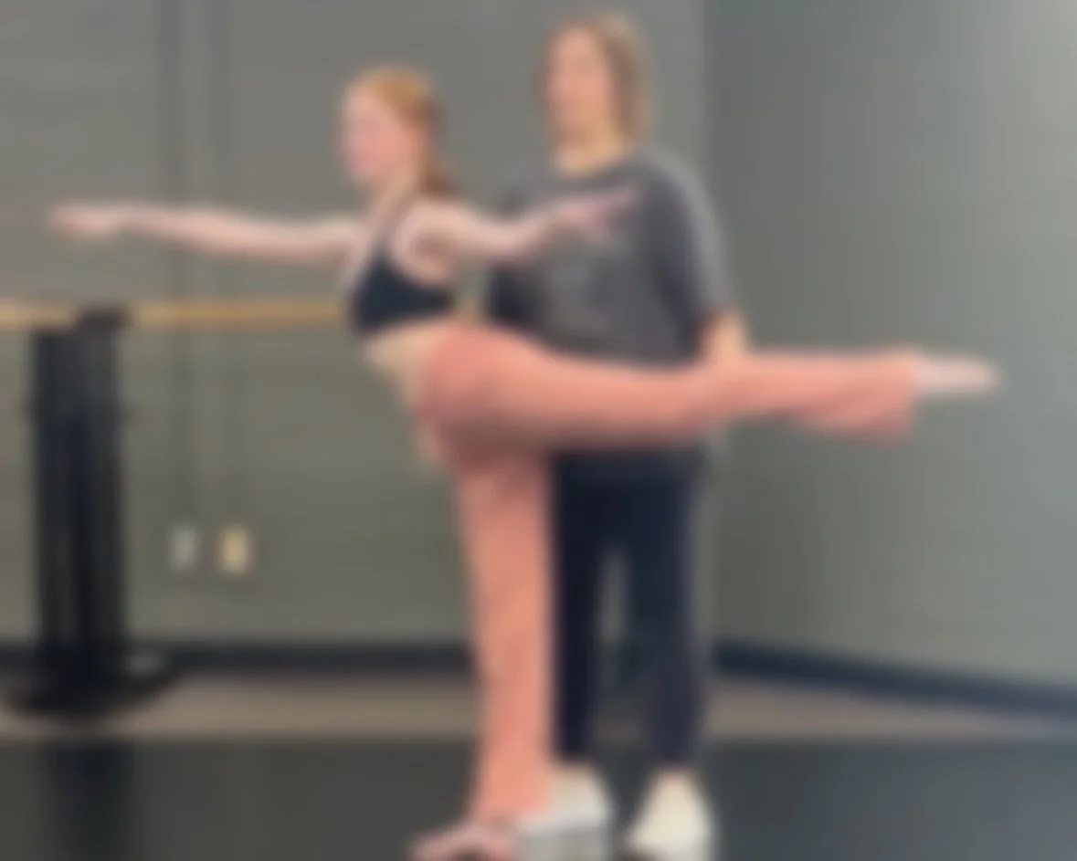 Two ballet dancers, one instructor and one student, practicing at a ballet barre in a dance studio.