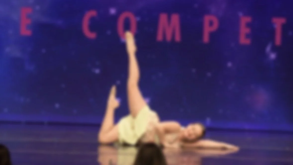 A dancer lying on the stage with one leg extended vertically, in front of a cosmic-themed backdrop with the words 'E C O M P E T' in red.