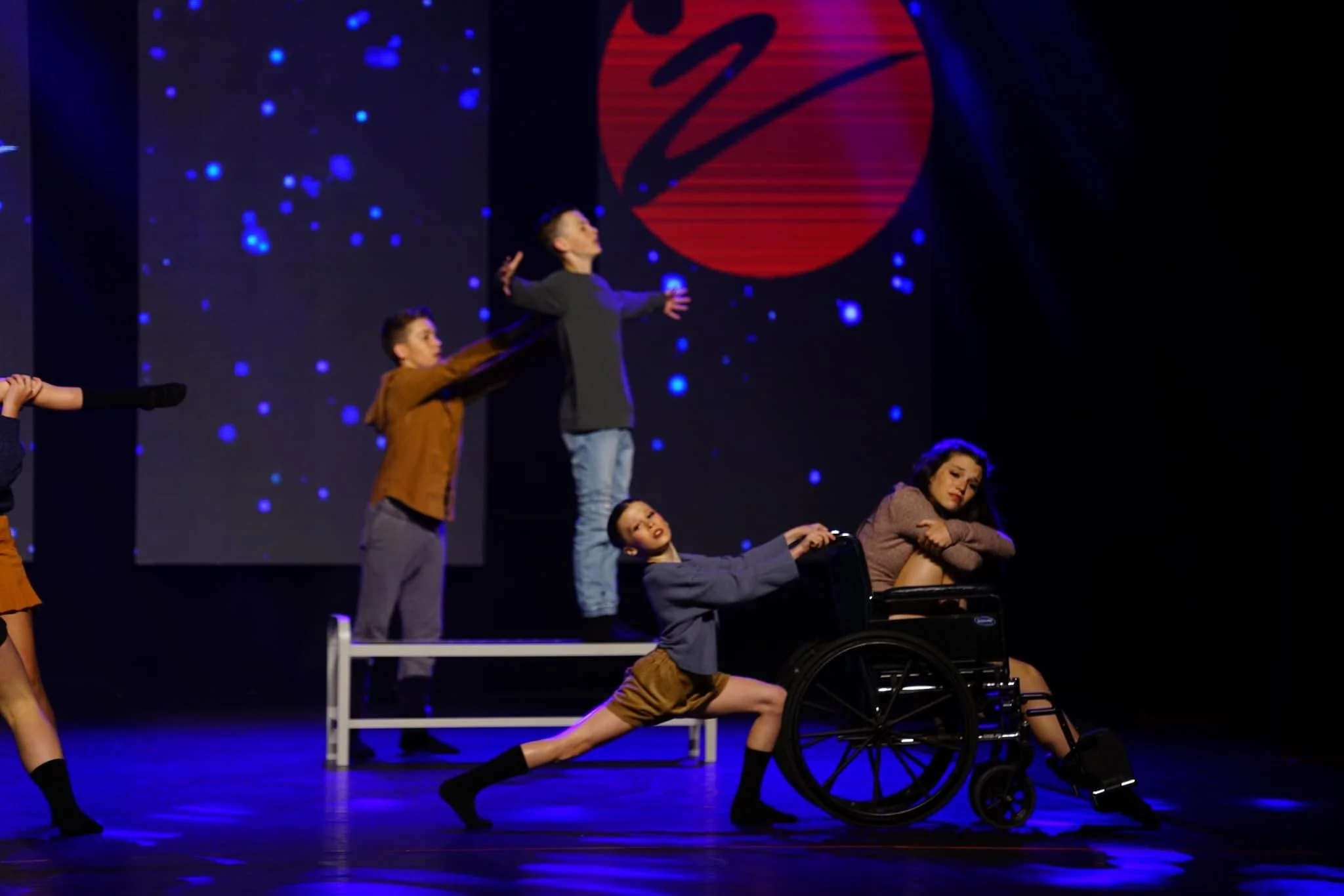 Stage performance featuring children, including one in a wheelchair, with a backdrop of space-themed visuals and a large red circle with a black signature-like mark.