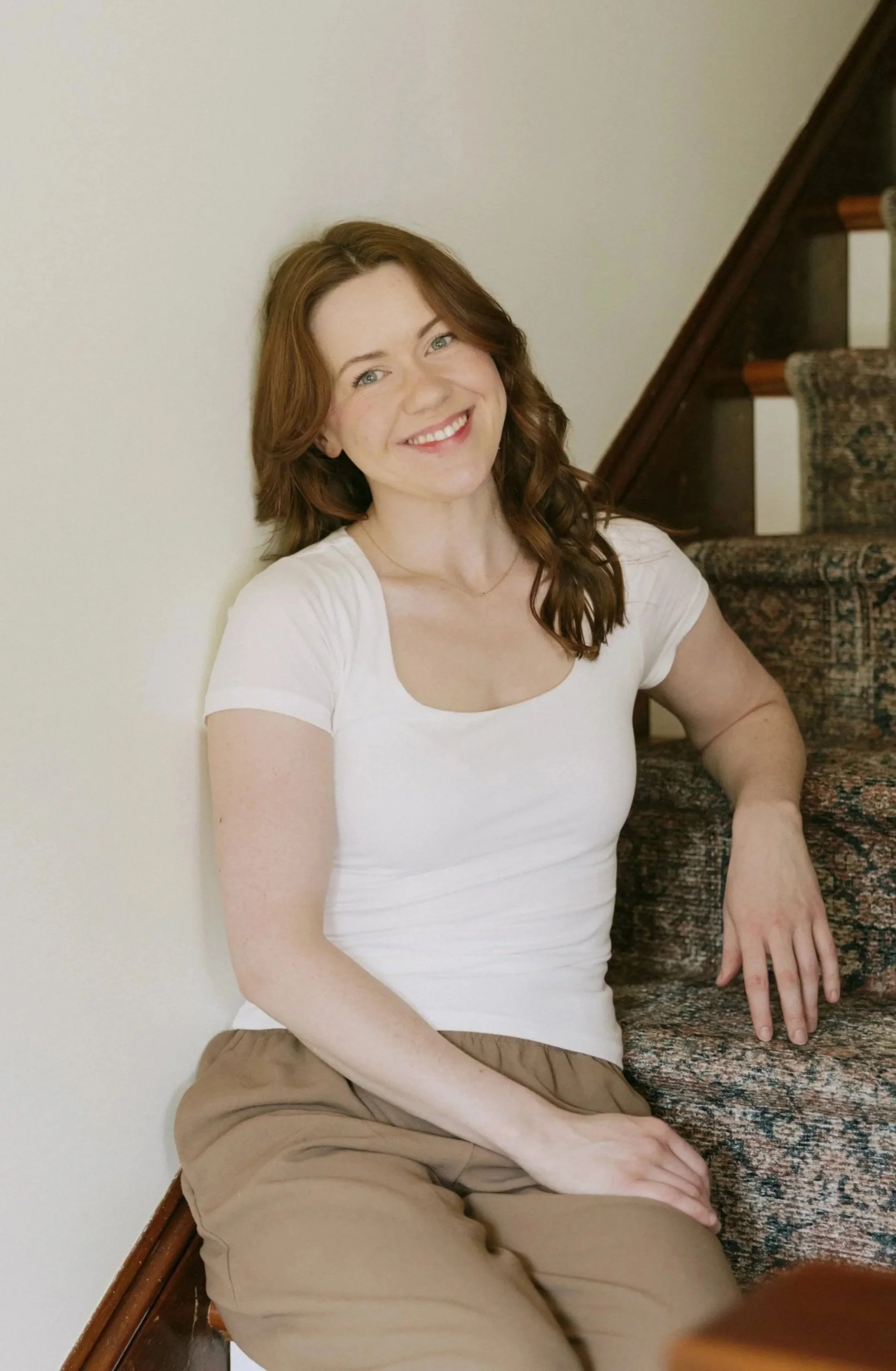 A young woman with red hair, wearing a white t-shirt and tan pants, sitting on a stairway with a smile, against a plain white wall.