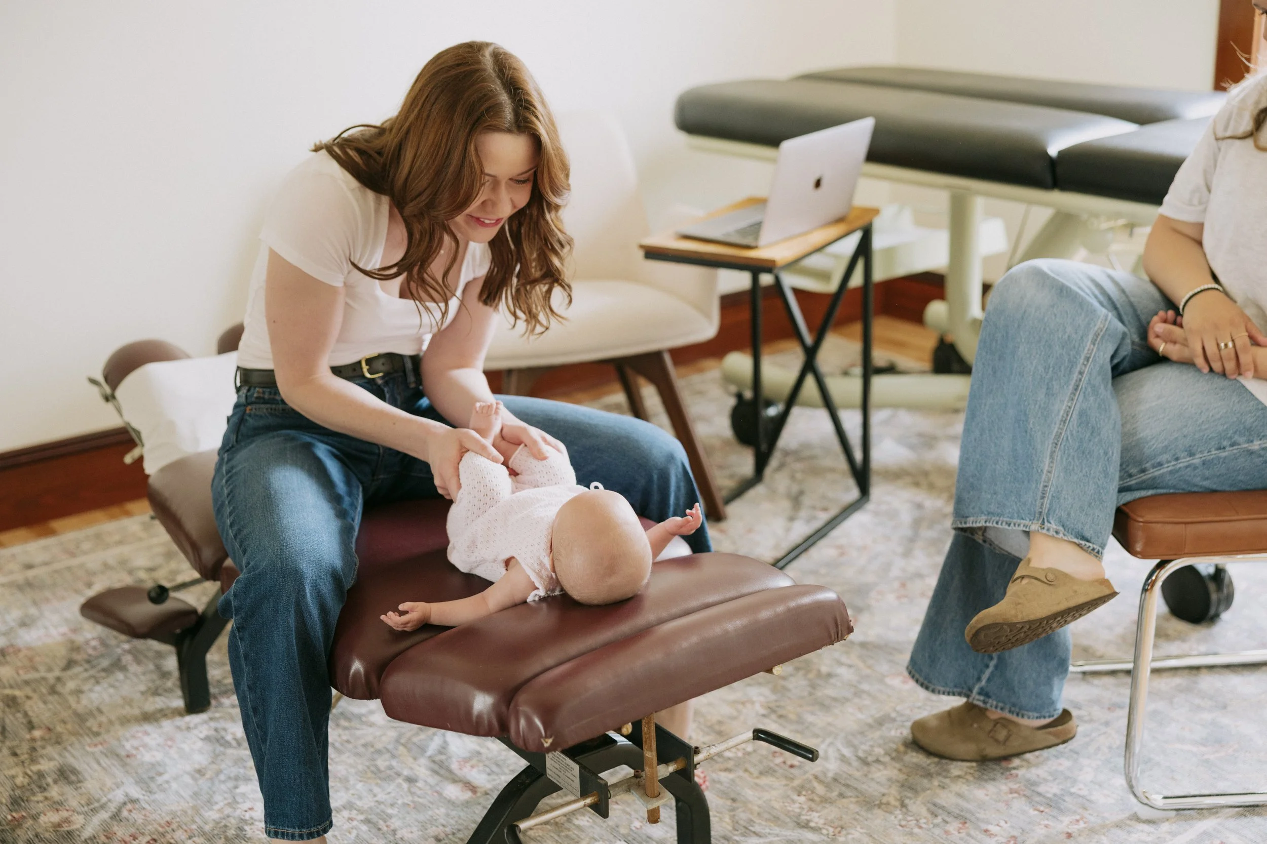 A young girl with brown hair smiling as she holds the feet of a baby lying on an examination table. The baby is dressed in a white outfit. In the background, there is a doctor with a stethoscope and medical equipment.