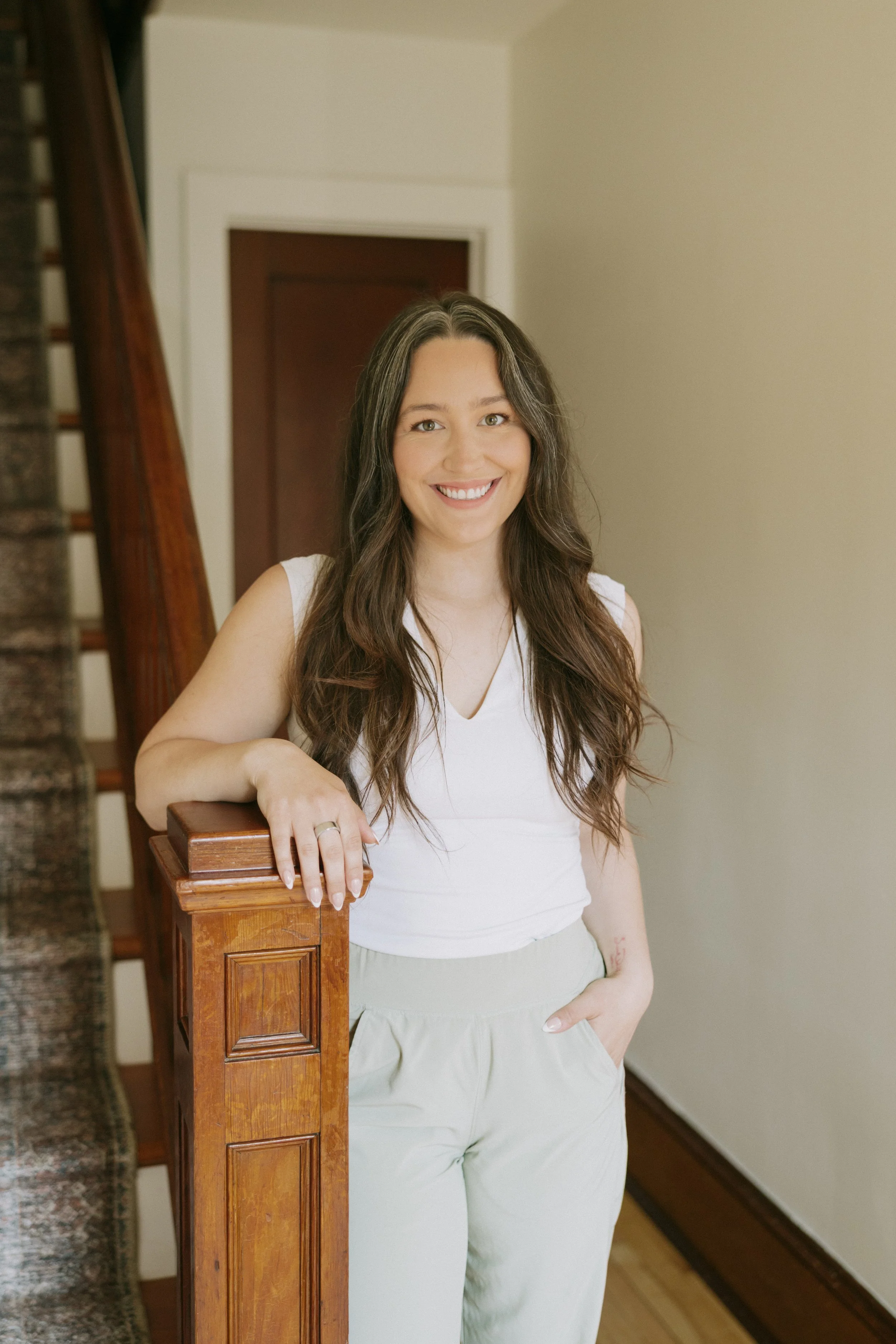 A woman with long, wavy brown hair and a bright smile, wearing a white sleeveless top and beige pants, standing outdoors by a wooden staircase railing in a home interior.