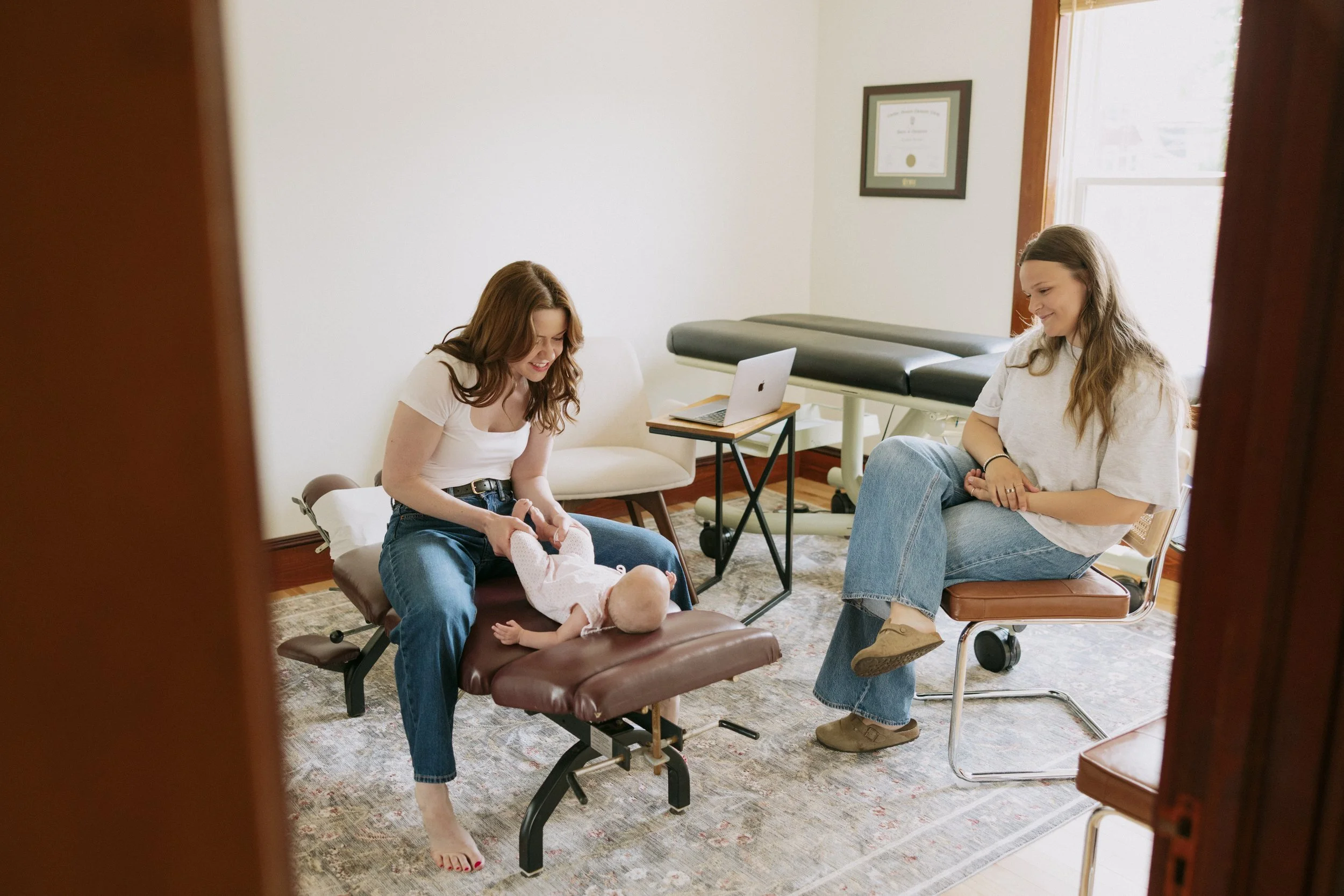 Two women in a medical setting with natural light, a framed diploma on the wall, a laptop, and a medical examination table. One woman is sitting on a massage table holding a baby, and the other woman is seated on a chair with her legs crossed, smiling.
