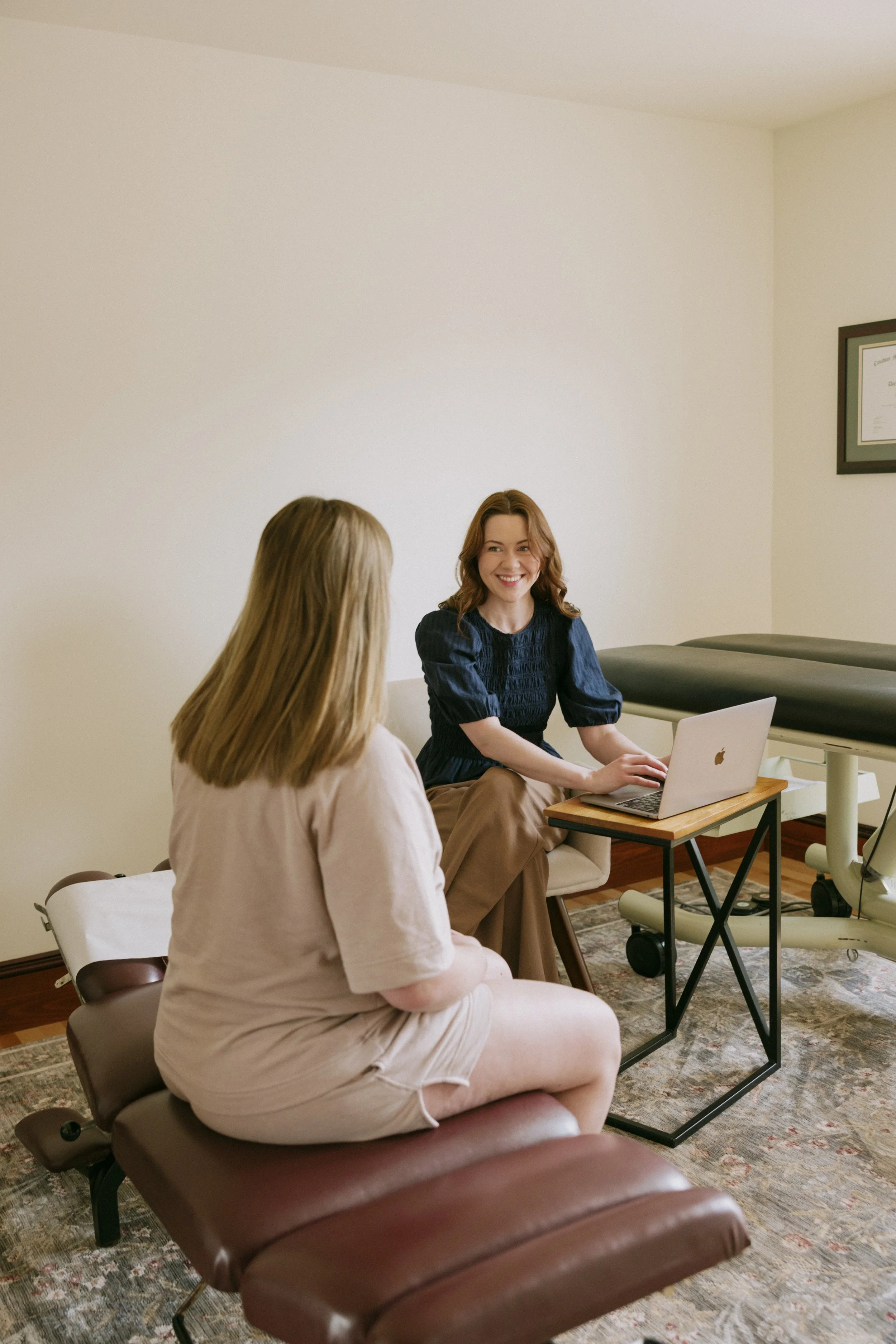 Two women having a conversation in an office, one with long blonde hair facing away, and the other with brown hair smiling, sitting in front of her laptop, with a treadmill and framed certificate on the wall in the background.