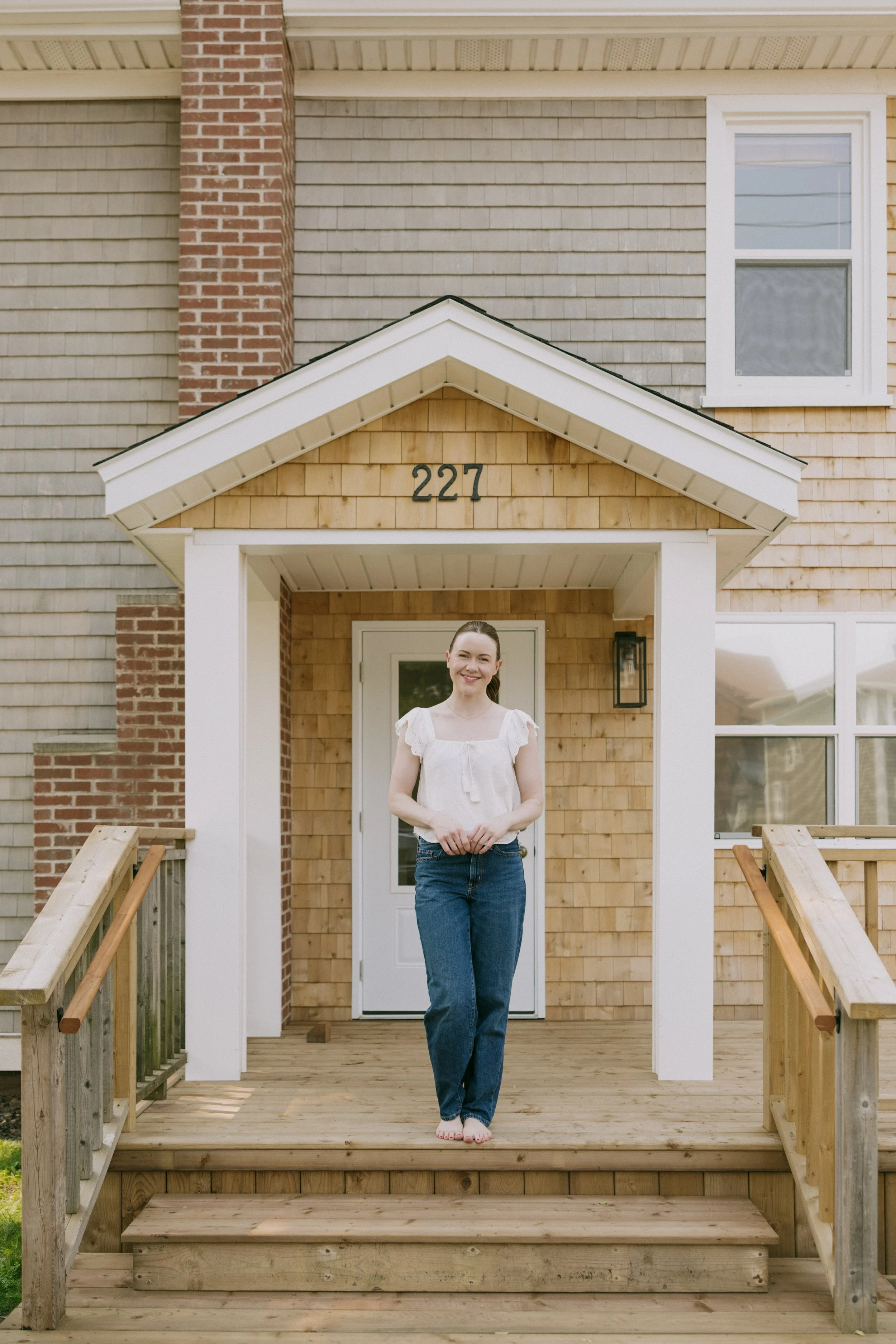 A woman standing barefoot on a wooden front porch stairs of a house, smiling, wearing a white blouse and blue jeans.