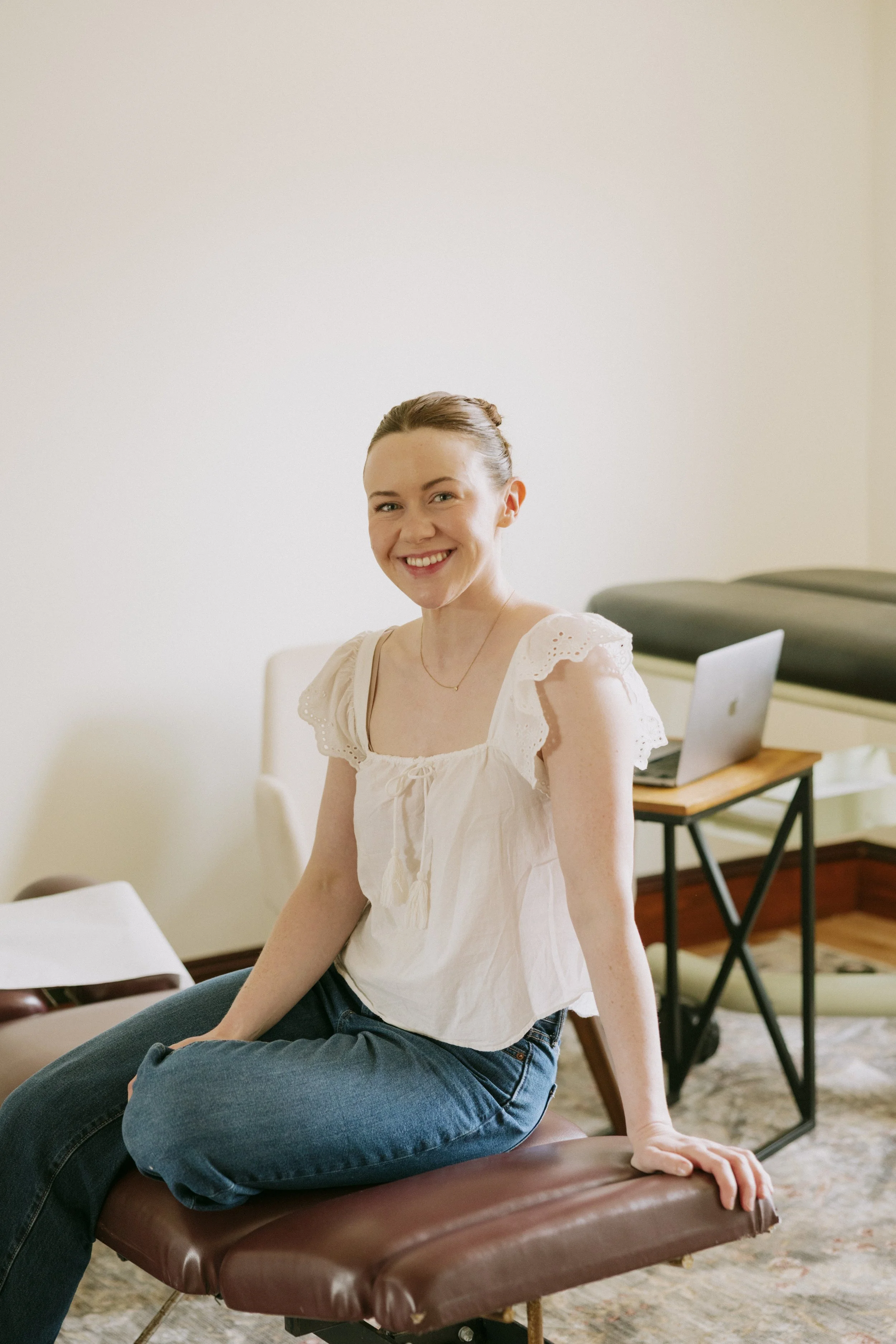 A young woman sitting on a massage table in a room with a laptop on a table behind her, smiling.