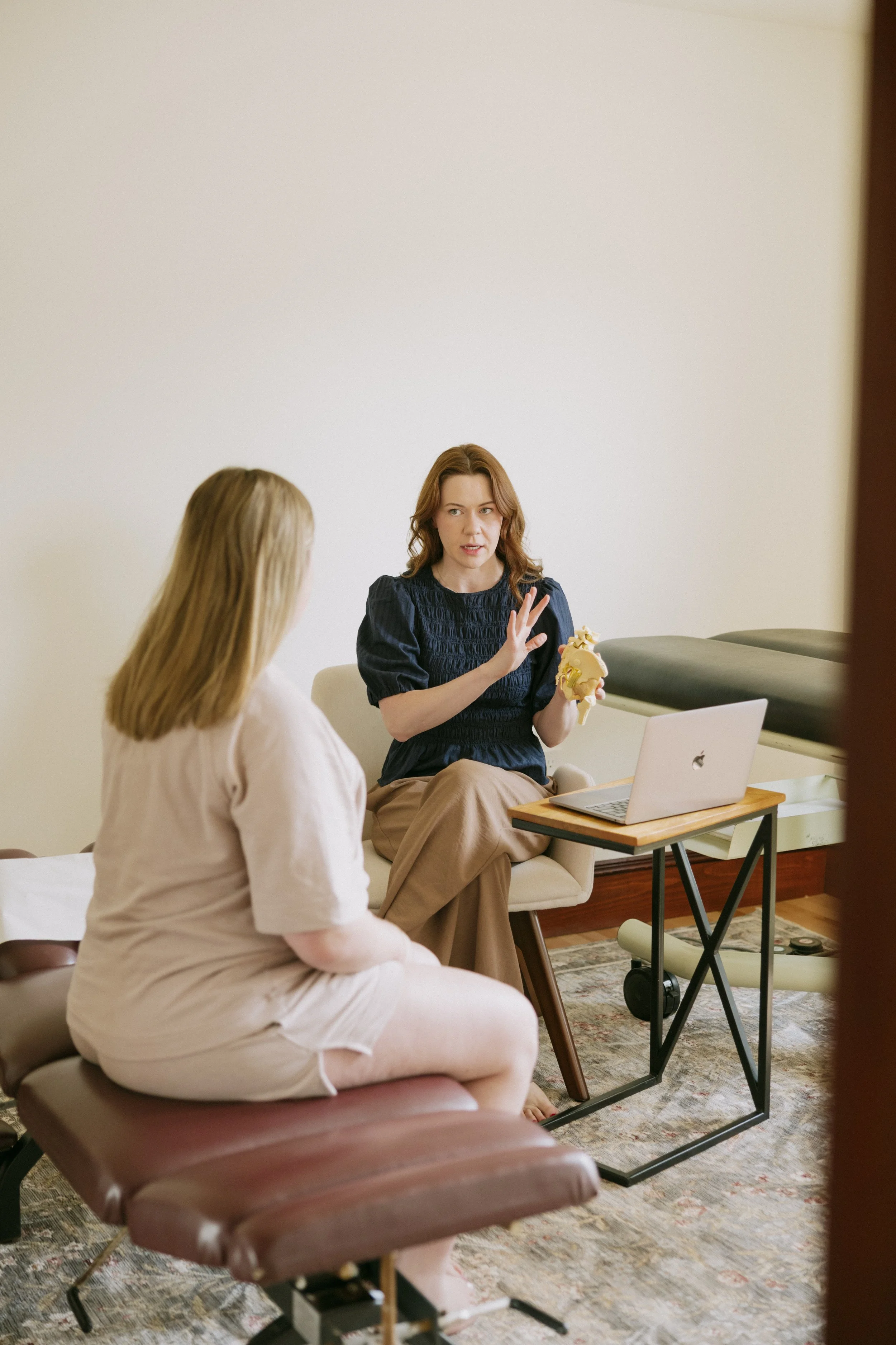 Two women are sitting in a room having a discussion. One woman has long blond hair and is facing away. The other woman has medium curly brown hair, is dressed in a navy blue top, and is gesturing with her right hand while holding a model of a human skull in her left hand. A laptop is open on a small table in front of them. The room has a neutral-toned wall, a patterned rug, and some furniture including a brown cushioned chair and a beige couch.