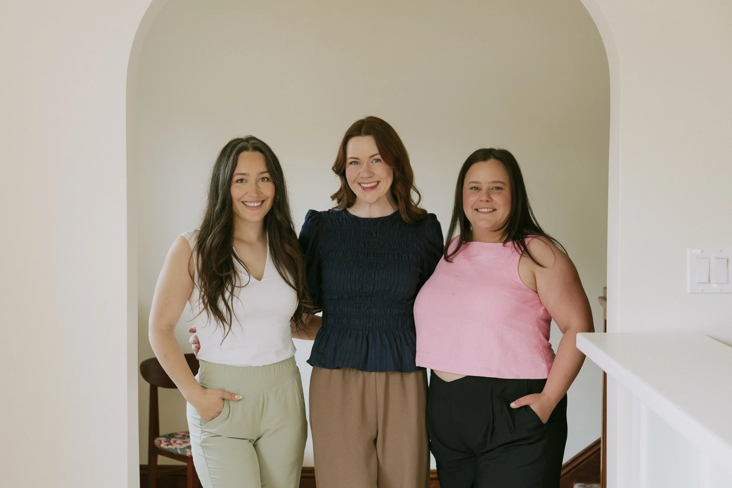 Three women standing close together indoors, smiling at the camera. The woman on the left has long, wavy dark hair and is wearing a white sleeveless top and light beige pants. The woman in the middle has shoulder-length red hair and is wearing a navy blue ruffled top and beige pants. The woman on the right has long straight dark hair and is wearing a pink sleeveless top and black pants.