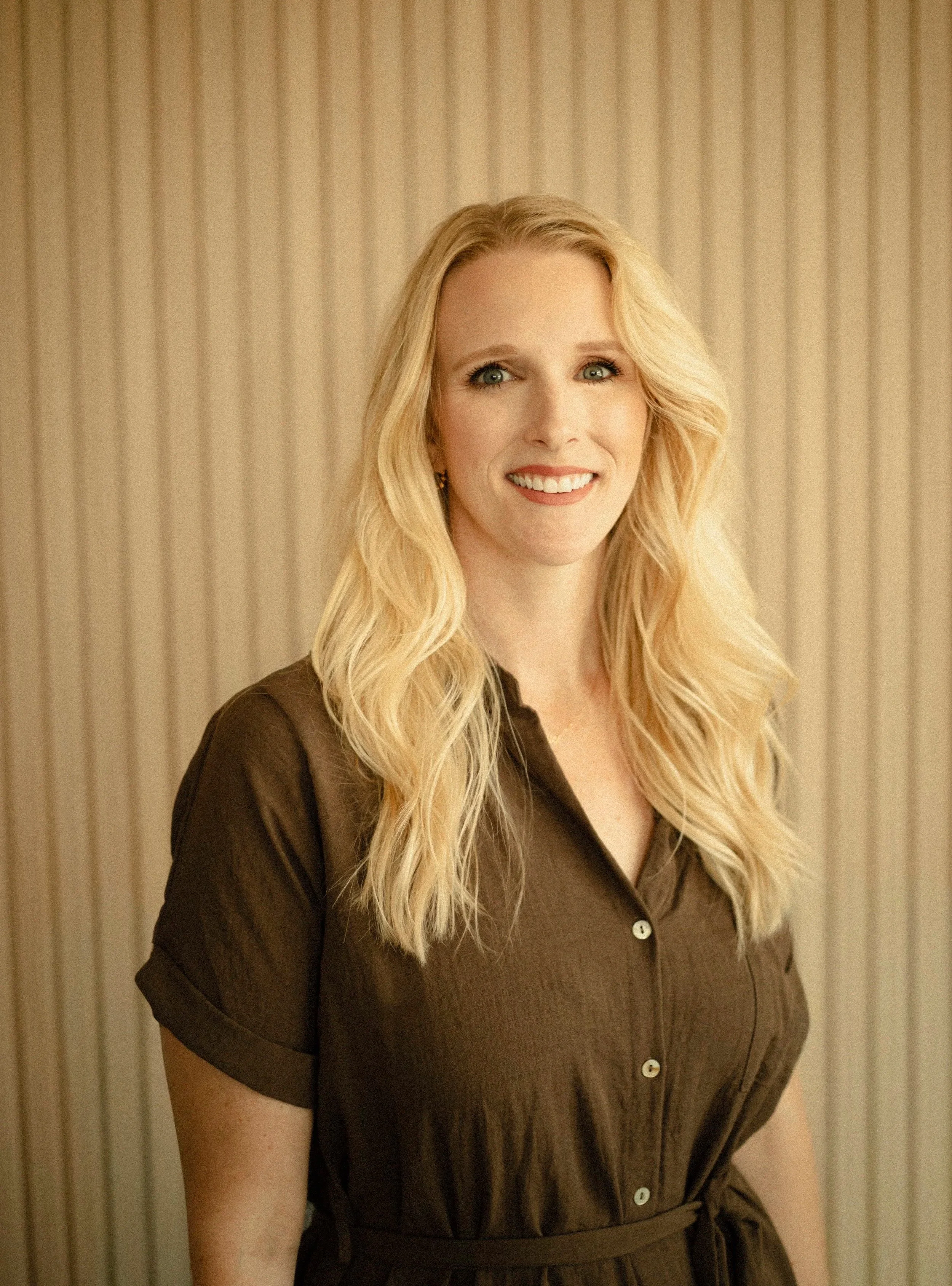 A woman with long blonde wavy hair wearing a brown dress with buttons and short sleeves, standing in front of a beige striped background, smiling.