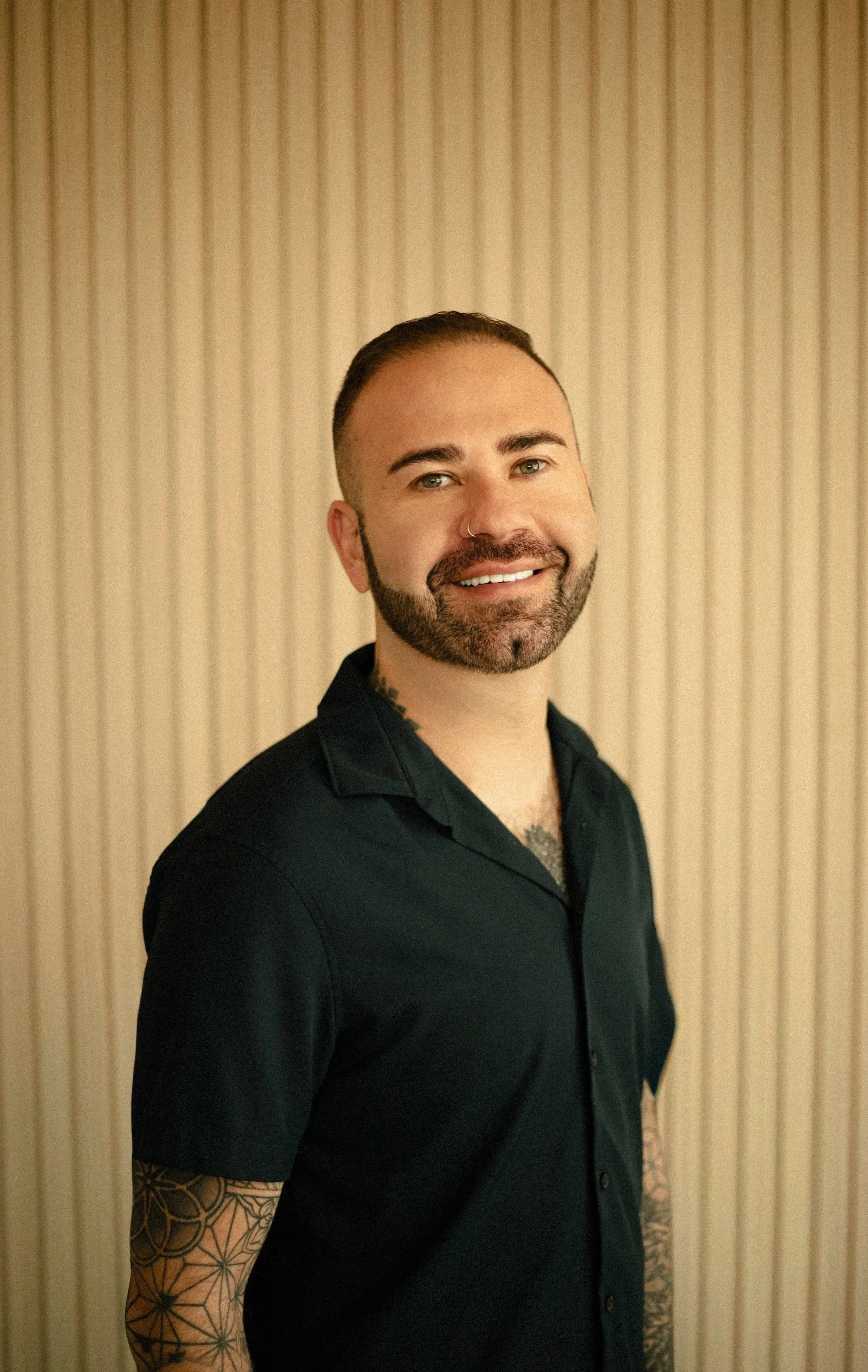 Portrait of a smiling man with a beard, mustache, and tattoos on his arms, wearing a black shirt, standing against a wood-paneled wall.