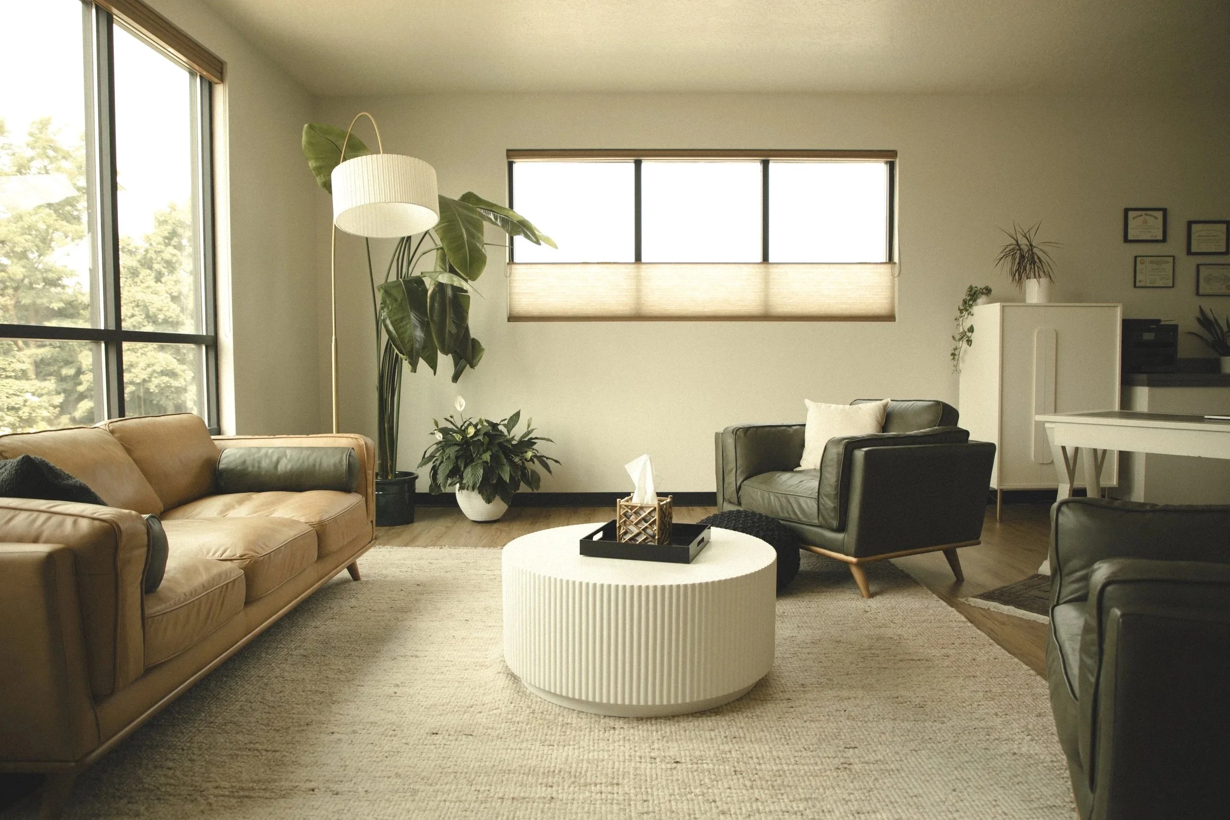Living room with a brown leather sofa, gray armchairs, a white textured round coffee table, houseplants, large windows, and framed certificates on the wall.