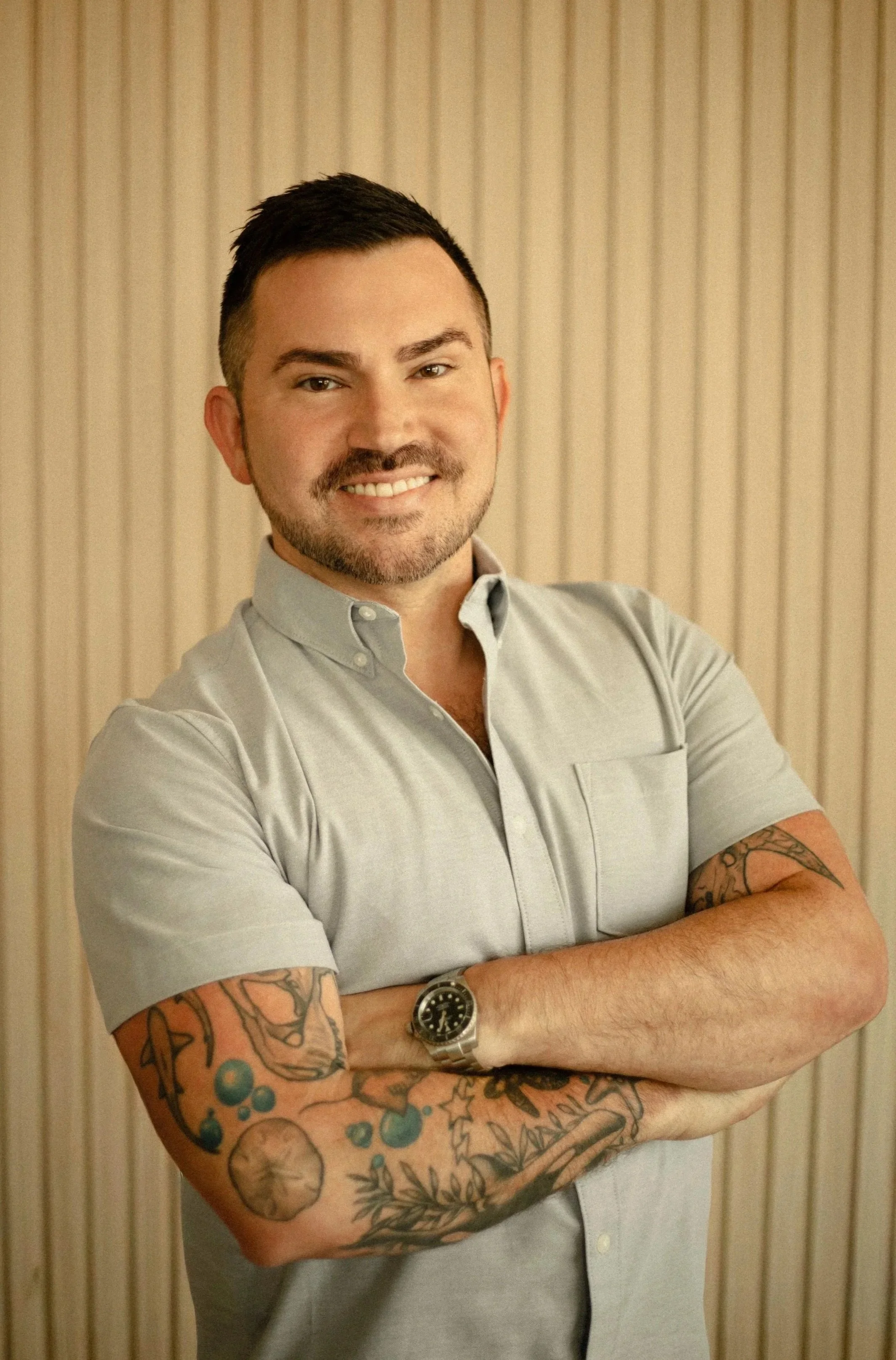 A man with short dark hair, a beard, and tattoos on his arms, wearing a gray button-up shirt with the sleeves rolled up, standing with arms crossed, smiling, and wearing a watch, in front of a beige striped wall.