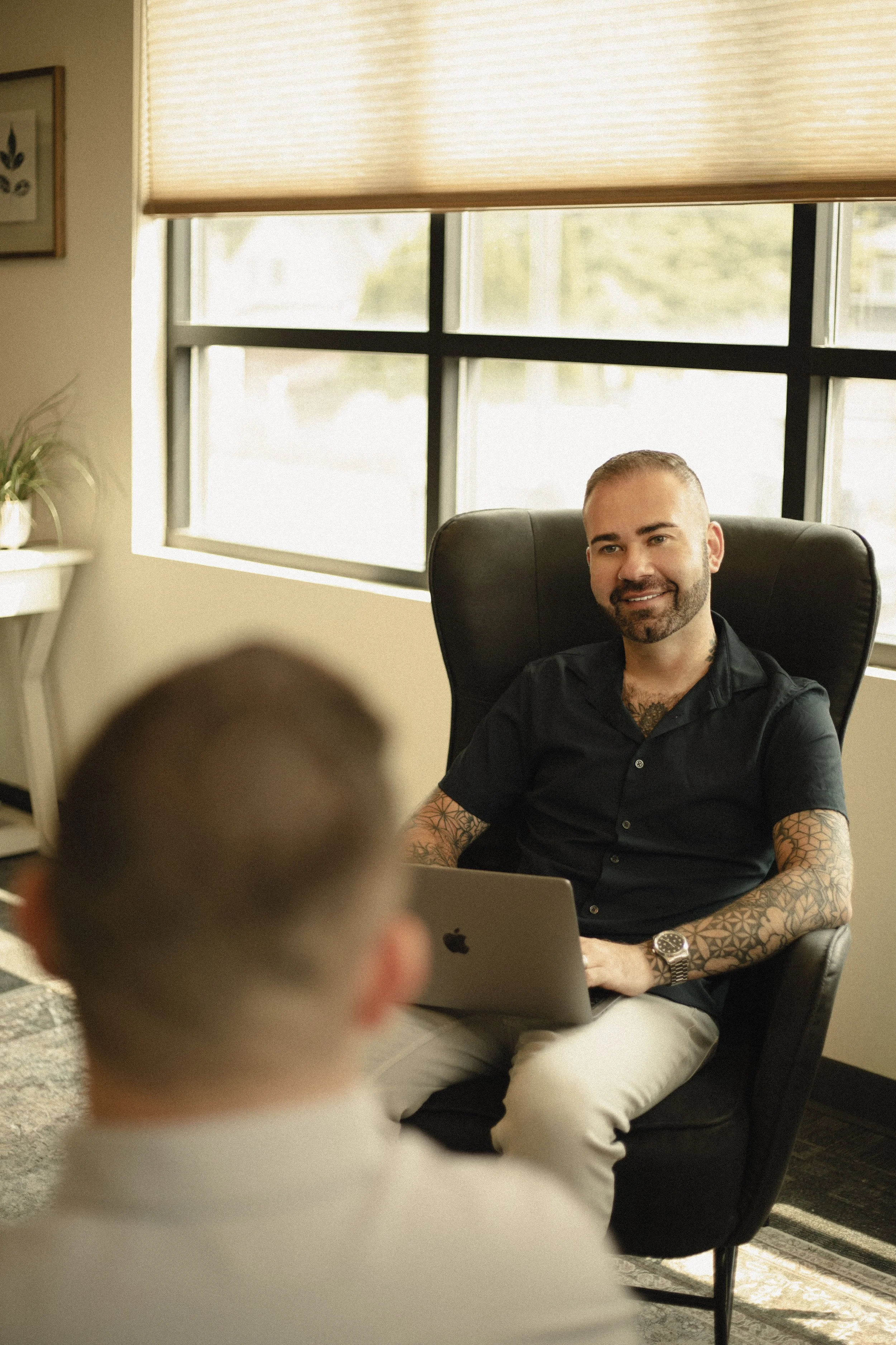 Man with tattoos smiling and sitting in a black office chair in a room with large window, beige blinds, a laptop, a watch, and a plant on a white shelf.