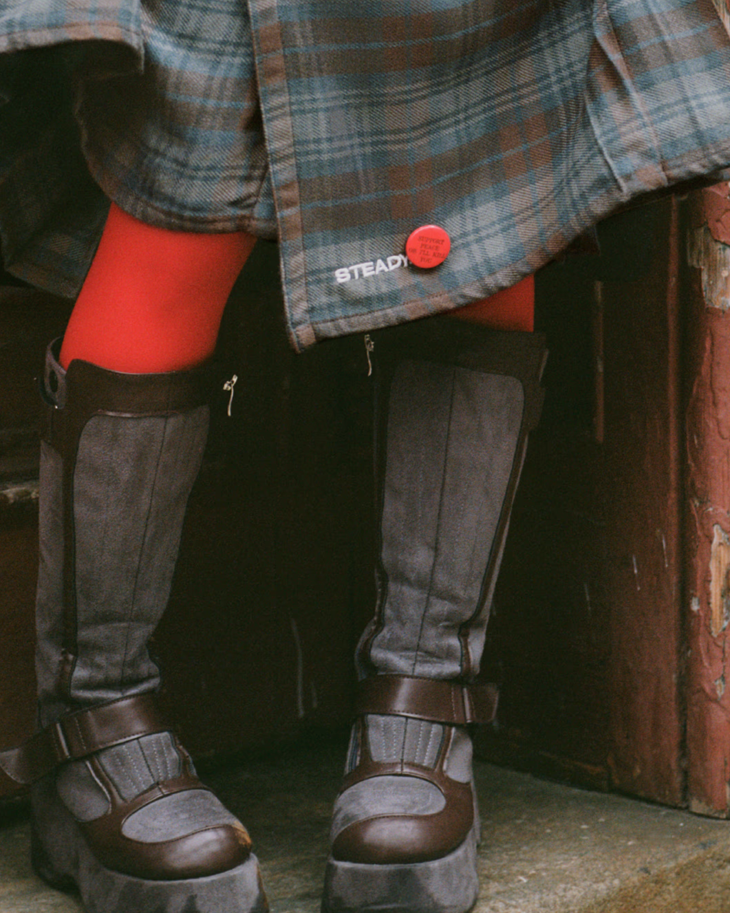 A person's lower body shows them wearing gray and black boots with brown straps, red tights, black pants, and a plaid shirt. A red pin is attached to the shirt, and the word "STEADY" is printed on the shirt.