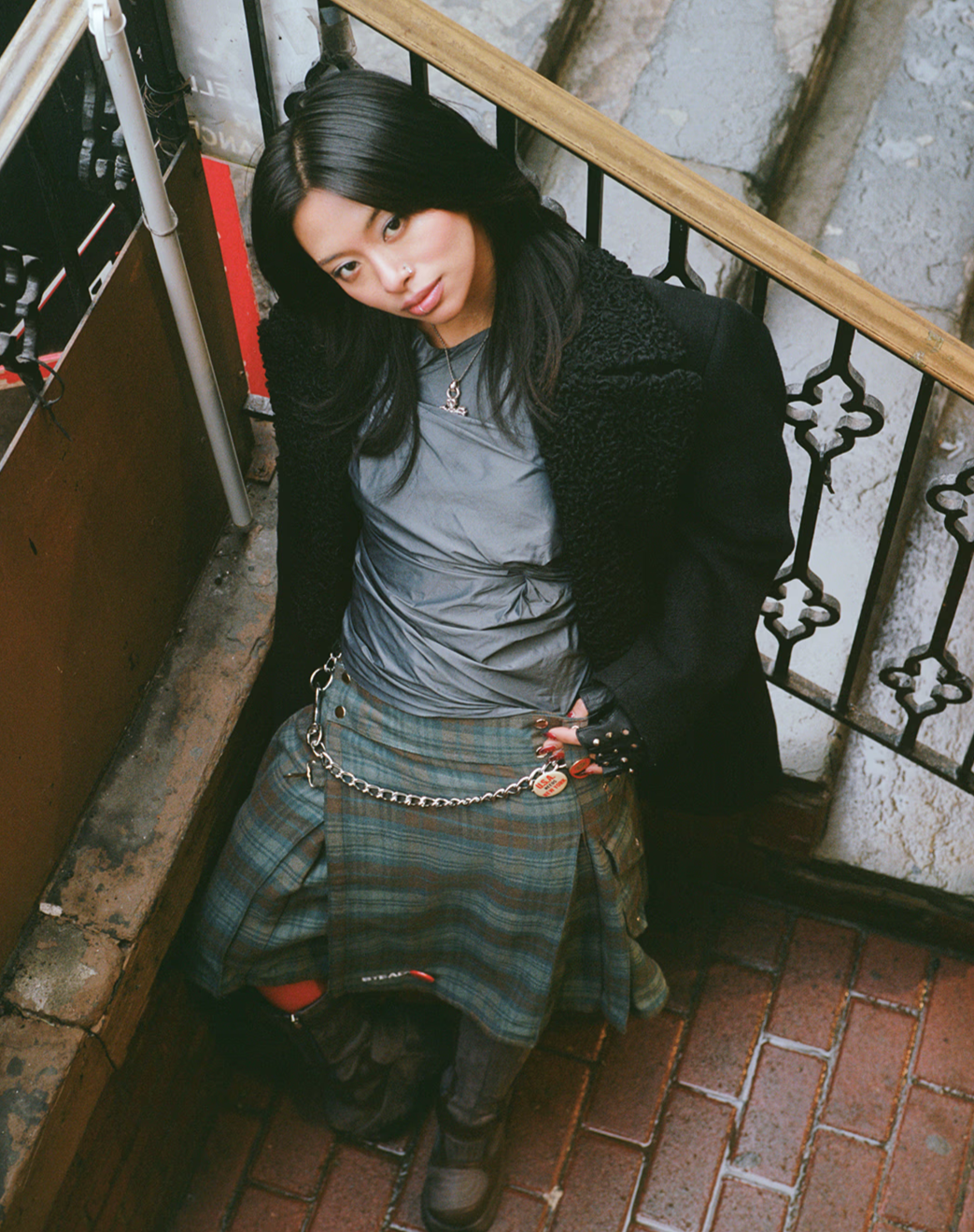 A young woman with long black hair, wearing a gray top, plaid skirt, and black jacket, sitting on a brick floor next to a metal railing, looking up at the camera.