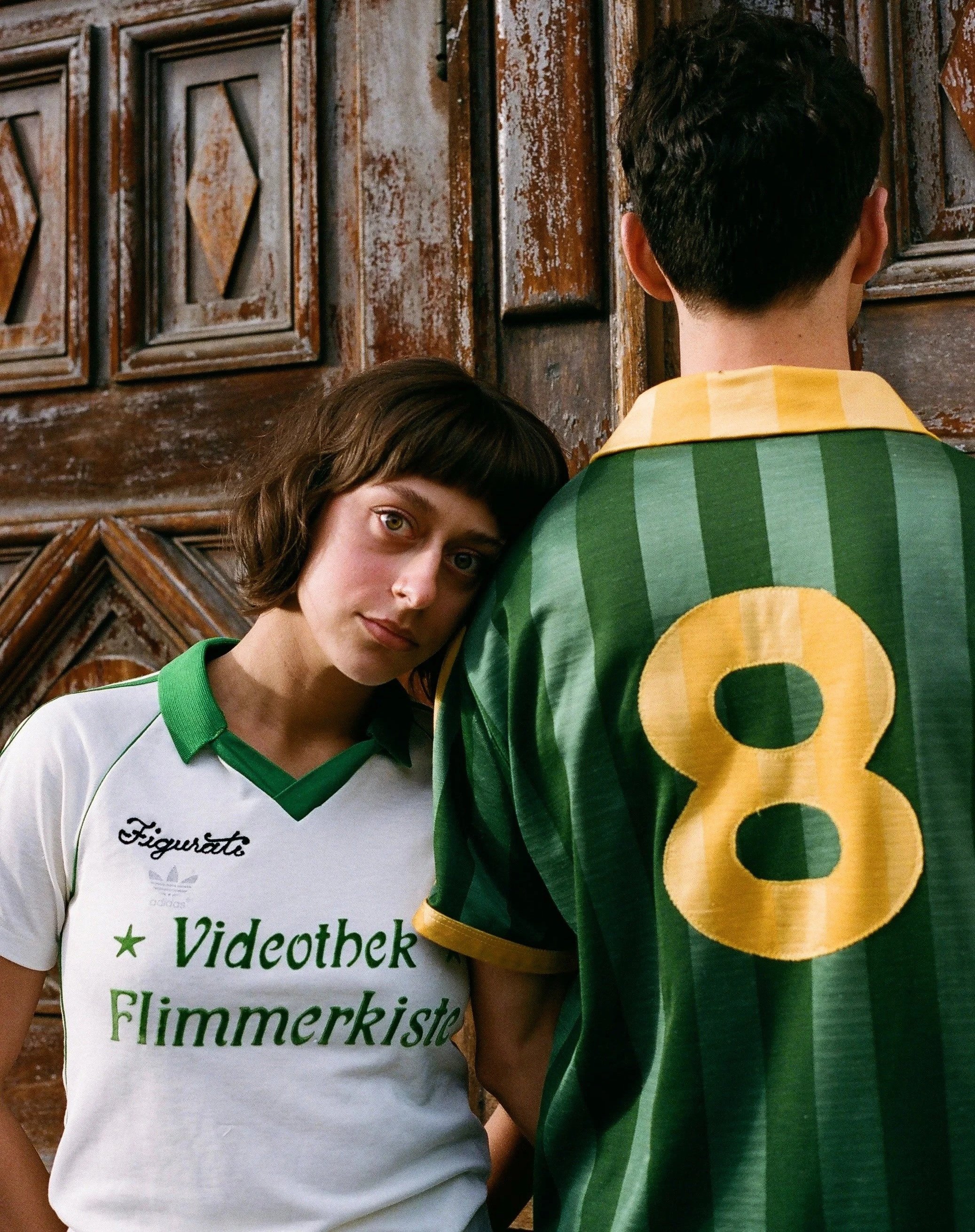 Close-up of two people standing closely together against a wooden background. The person on the left has light skin, short brown hair, and green eyes, and is wearing a white sports jersey with green accents and written text. The person on the right h