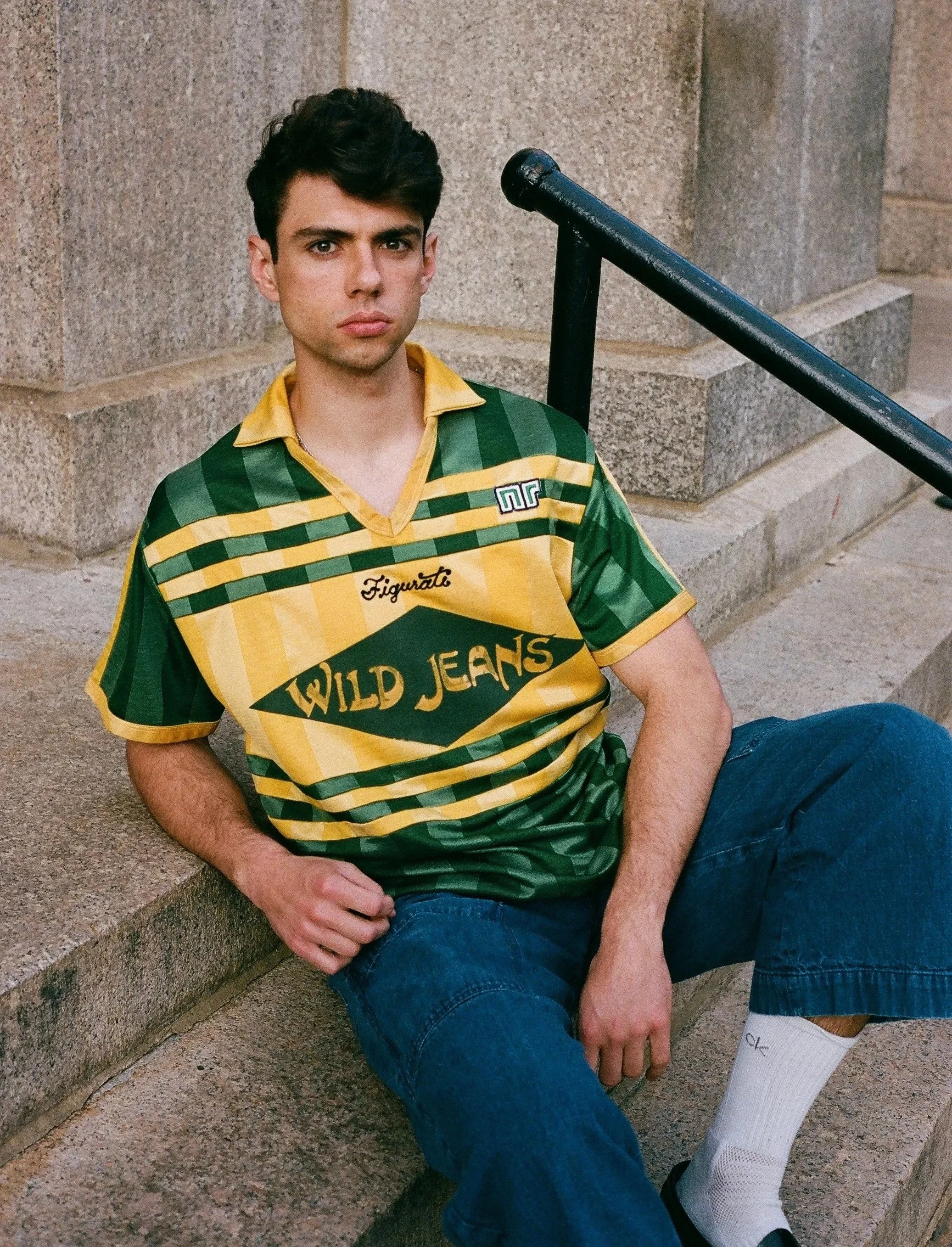 A young man with dark hair and light skin sitting on stairs outdoors, wearing a green and yellow striped rugby shirt with 'Wild Jeans' written on it, blue jeans, and white socks with black shoes. He has a serious expression, and a staircase handrail 