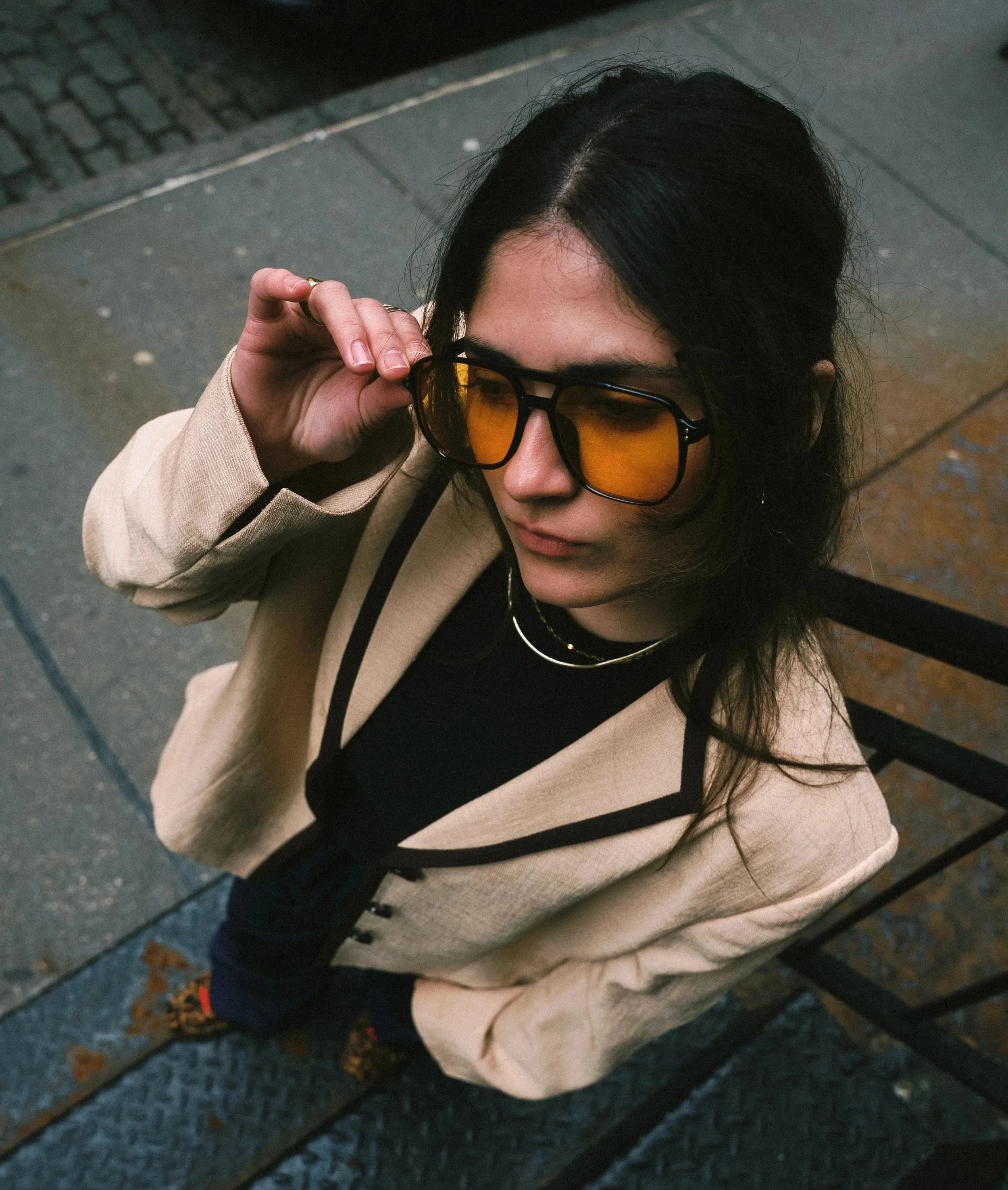 Person with long dark hair wearing sunglasses with orange lenses, a beige coat, and a black shirt, sitting on a bench outdoors, holding the sunglasses with their right hand, on a sidewalk with paving stones.