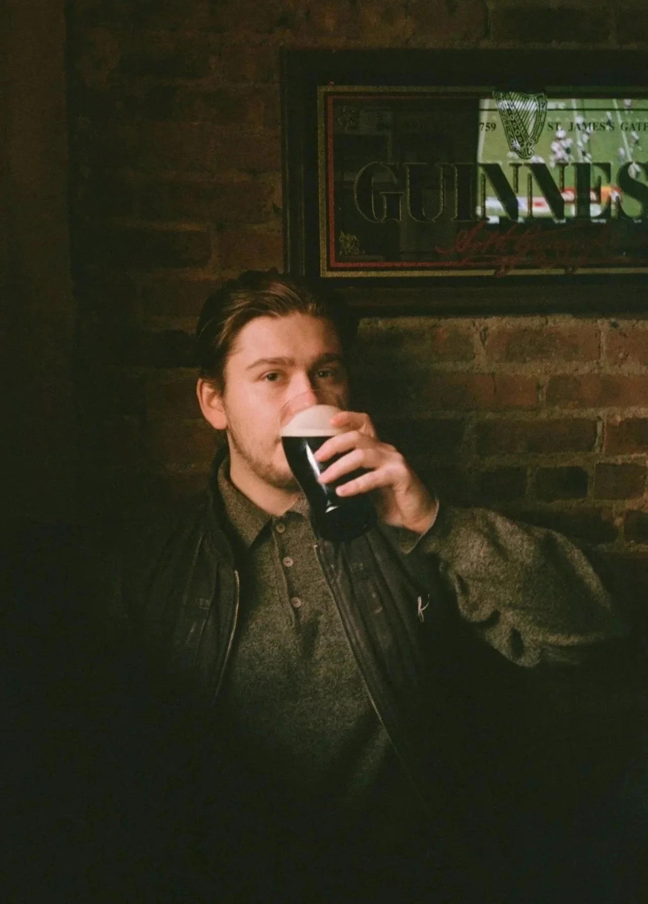 A young man with brown hair and facial hair sitting in a dimly lit pub, drinking a dark beer from a pint glass, against a brick wall with a Guinness sign.