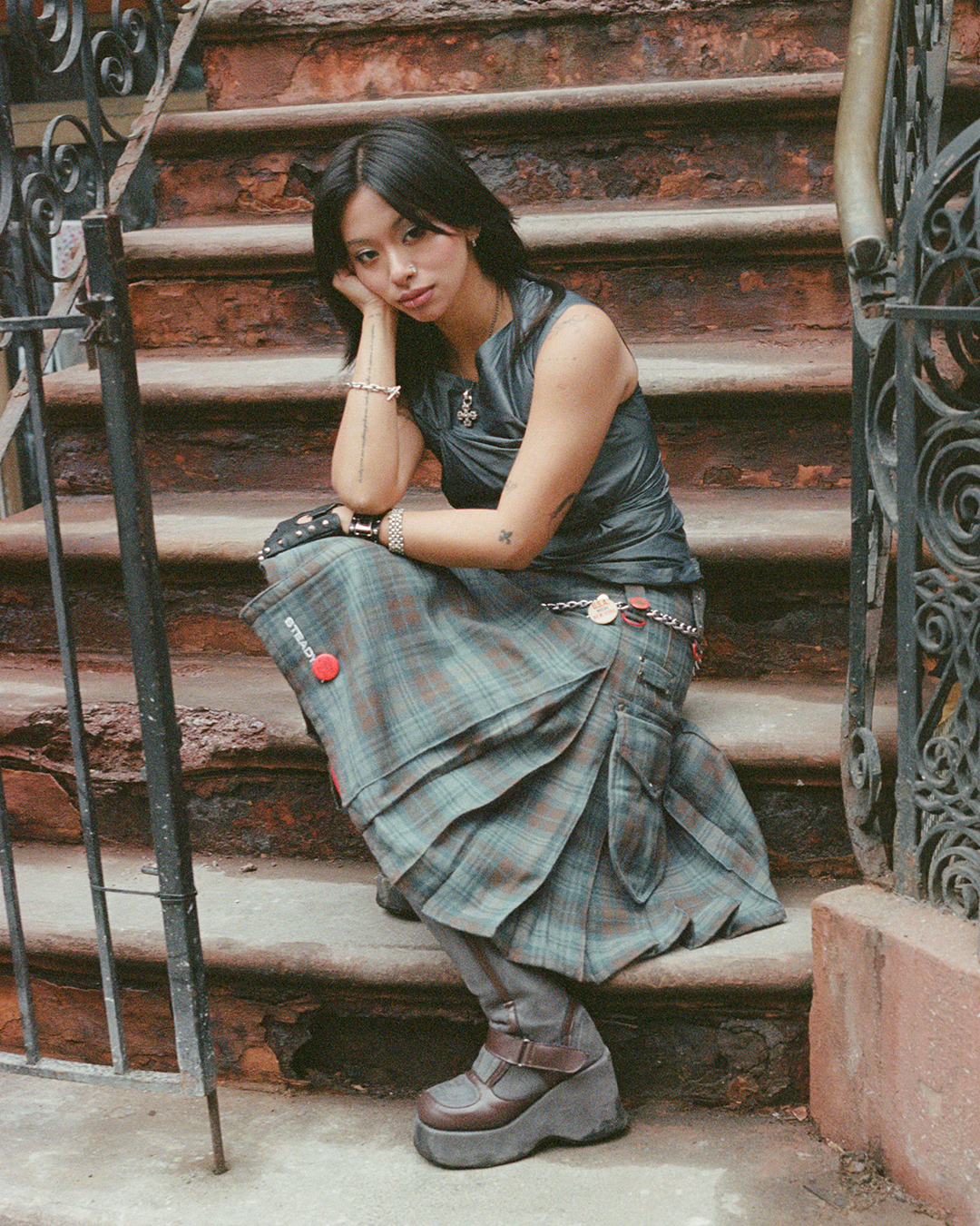 A young woman with black hair, wearing a black leather top, plaid skirt, and chunky shoes, sitting on outdoor stone steps beside a wrought iron railing, with a contemplative expression.