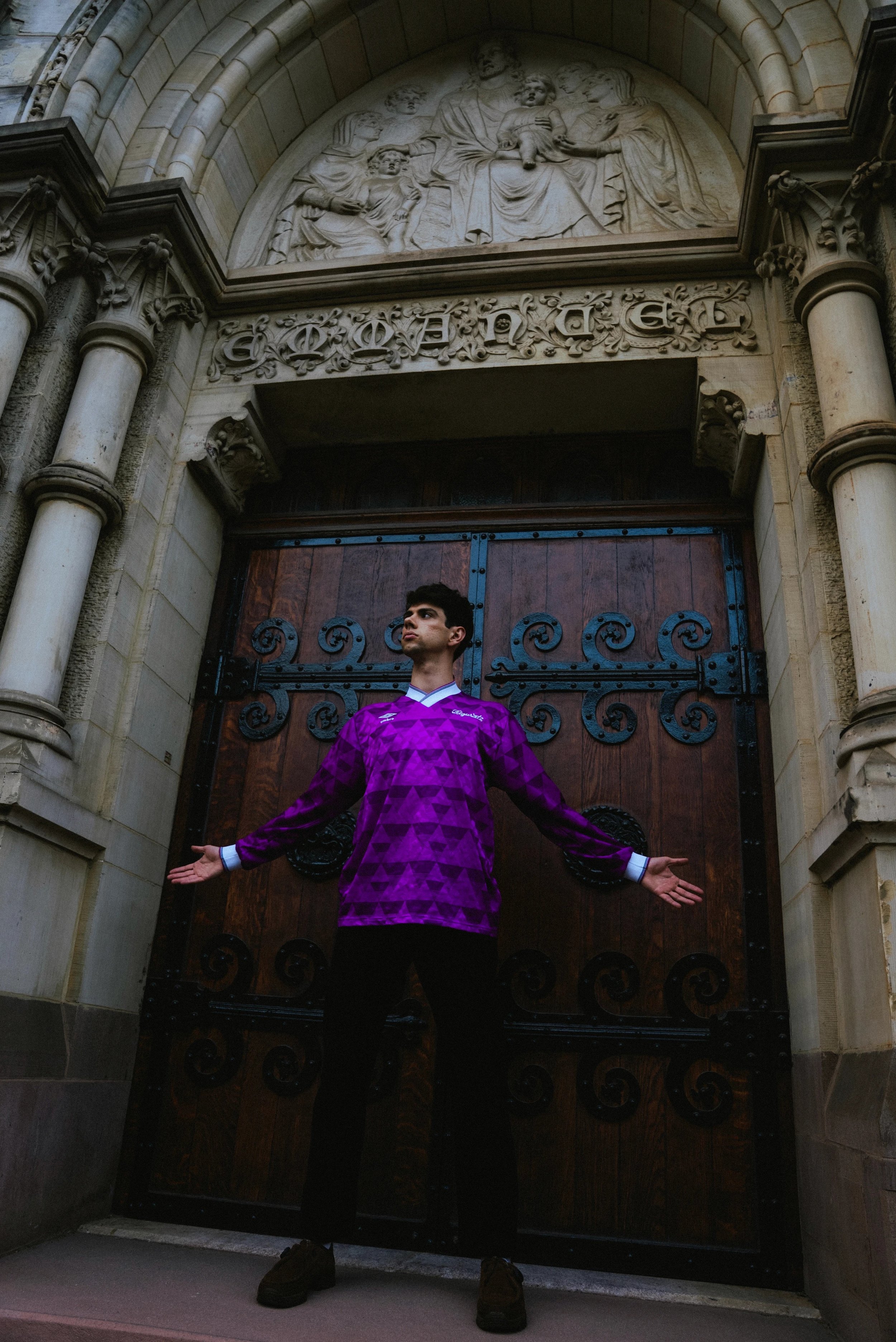 Young man in a purple geometric patterned shirt standing in front of a large wooden gate with iron scrollwork, under a stone arch with religious relief sculpture and decorative carvings.