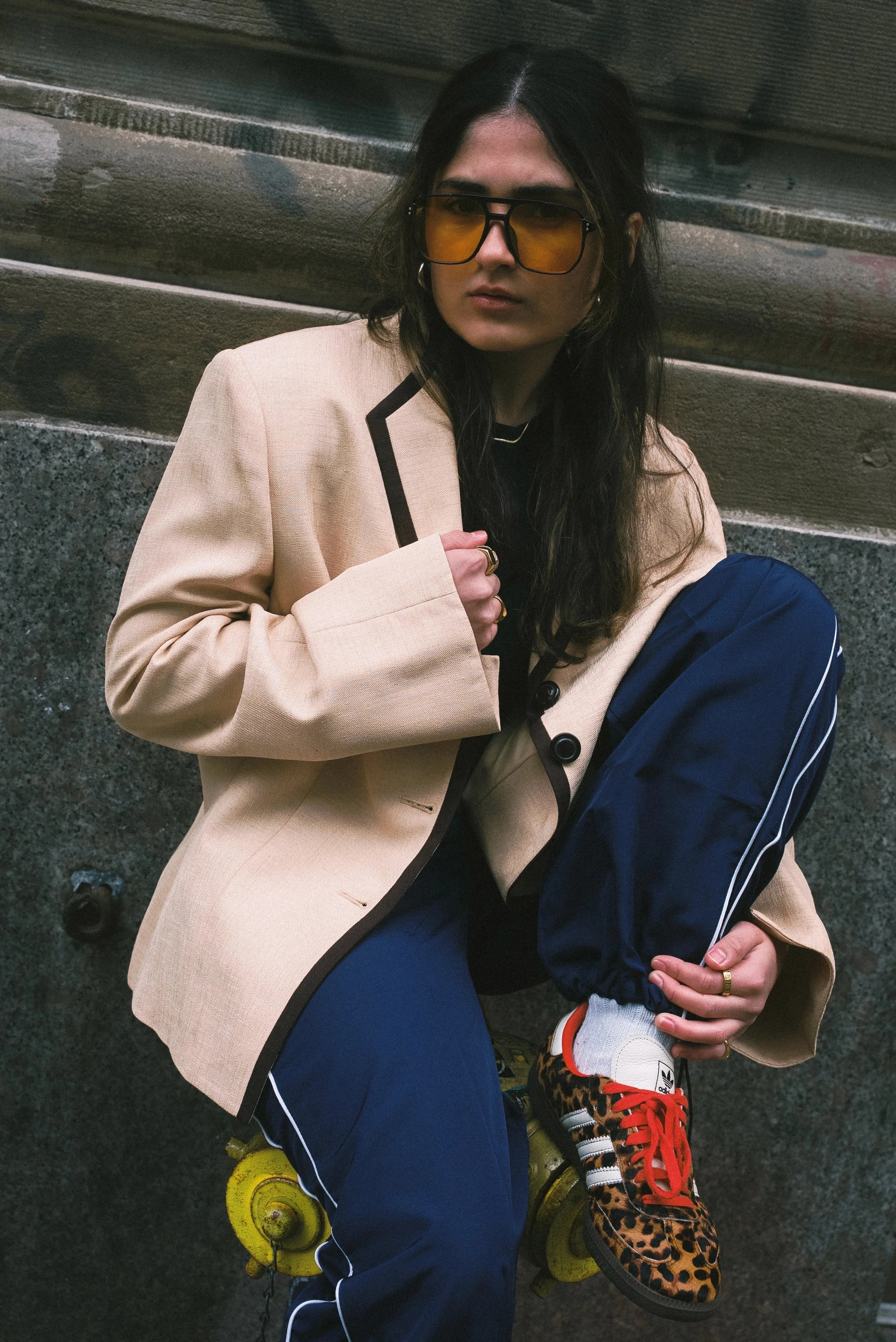 A woman wearing a beige blazer, black shirt, and yellow sunglasses, sitting on yellow roller skates with leopard print shoes, against a textured concrete wall.