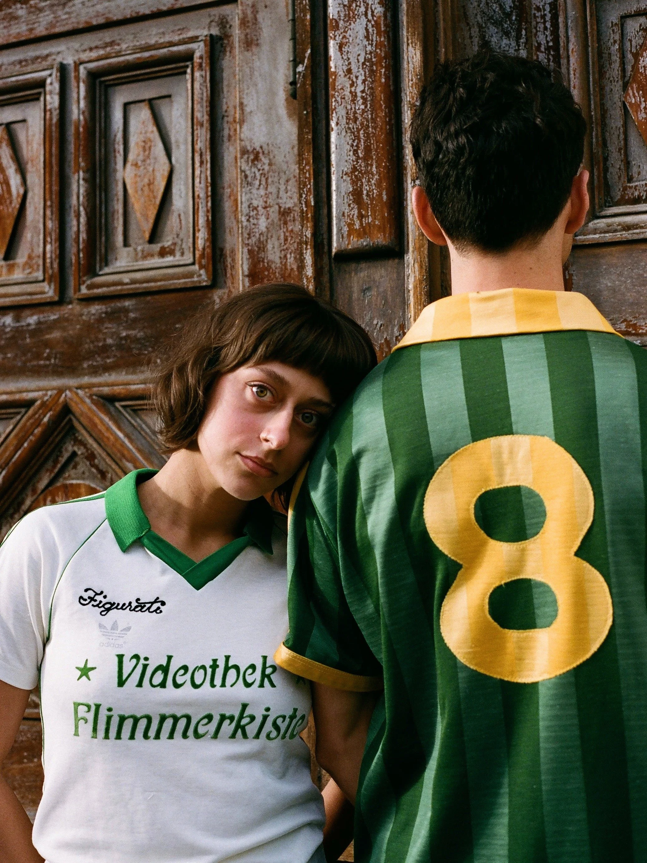 A young woman with short brown hair and a young man with dark, curly hair standing against a wooden wall. The woman is wearing a white soccer jersey with green accents and text. The man is wearing a green sports jersey with yellow accents and the number 8. The woman is leaning her head on the man's shoulder, and she is looking directly at the camera.