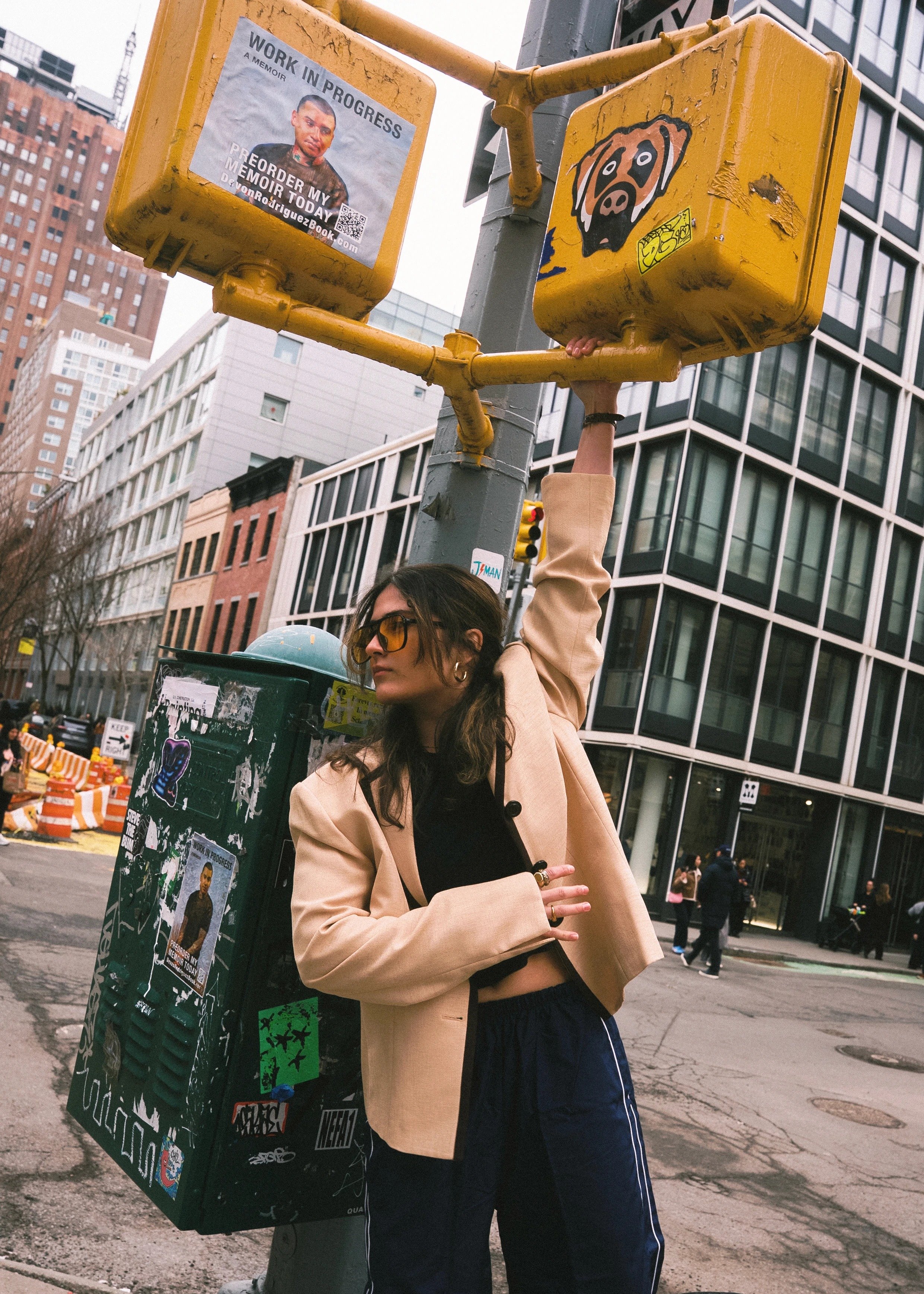 A person with glasses and long hair standing next to a utility pole, holding onto a yellow traffic signal box with both hands. 