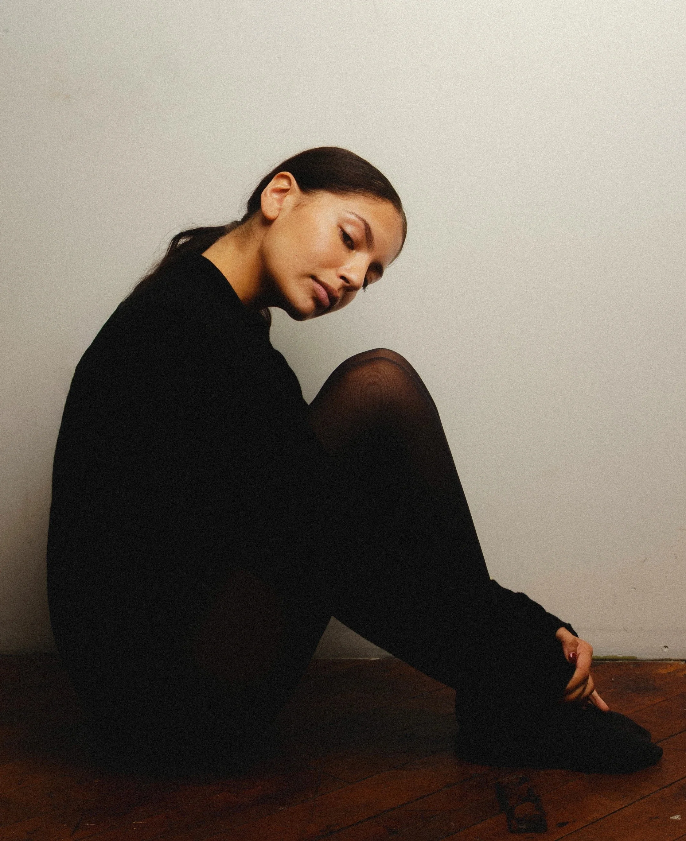 A woman sitting on a dark hardwood floor with her legs crossed, wearing black clothes and fishnet tights, against a plain light-colored wall.