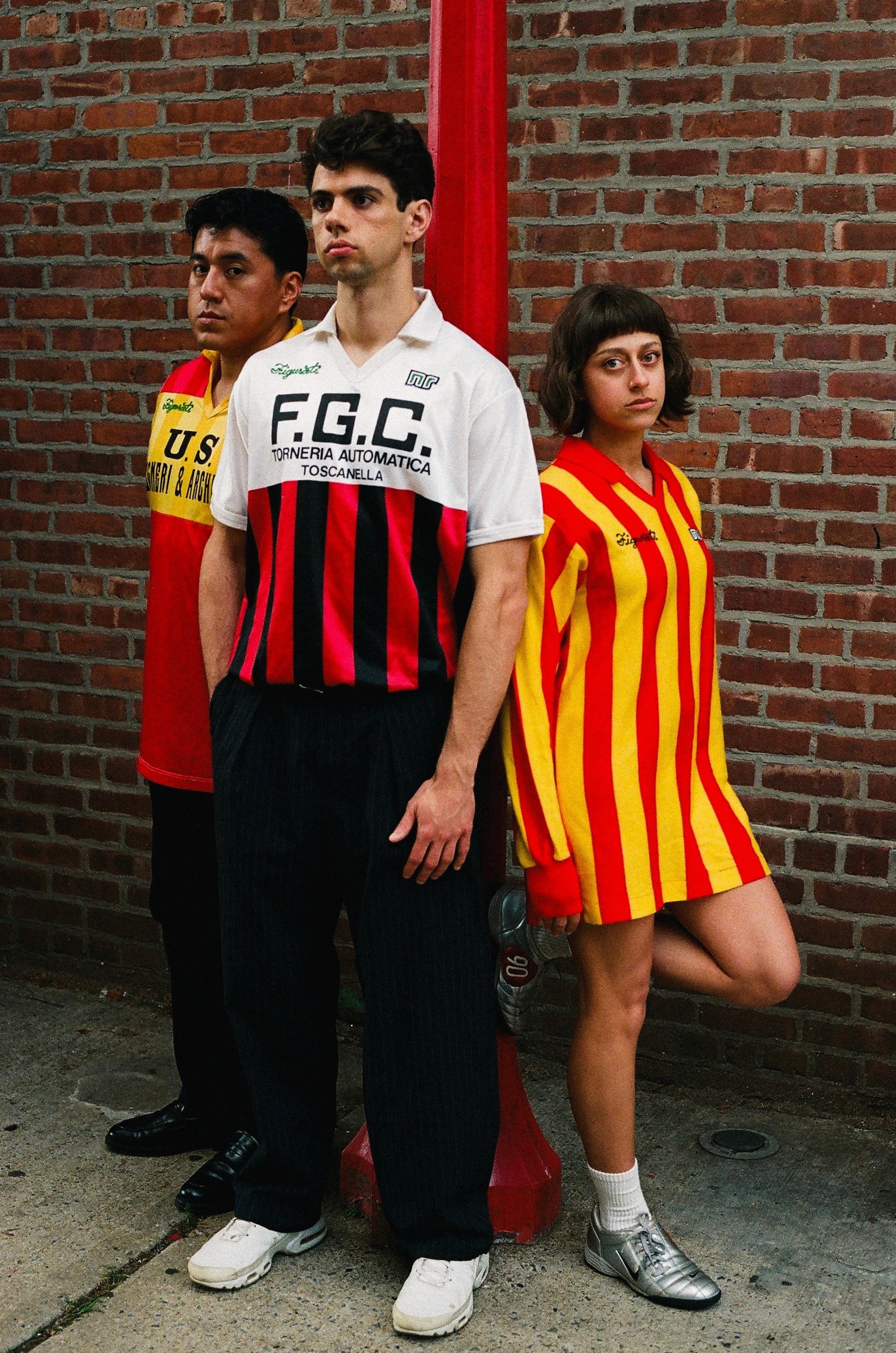 Three young adults standing against a brick wall with a red post. The person in the middle is a man with dark hair, wearing a white and red striped shirt with black pants and white sneakers. The person on the left is a man with dark hair, wearing a y
