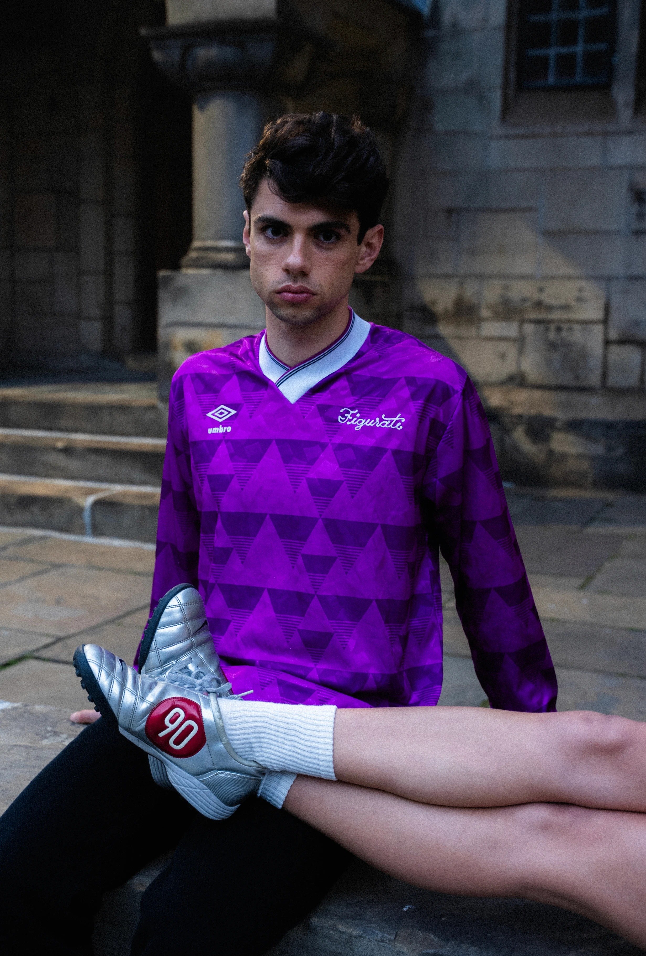 A soccer player in a purple uniform with football boots on his lap and looking at the camera, outside near a stone building with stairs.