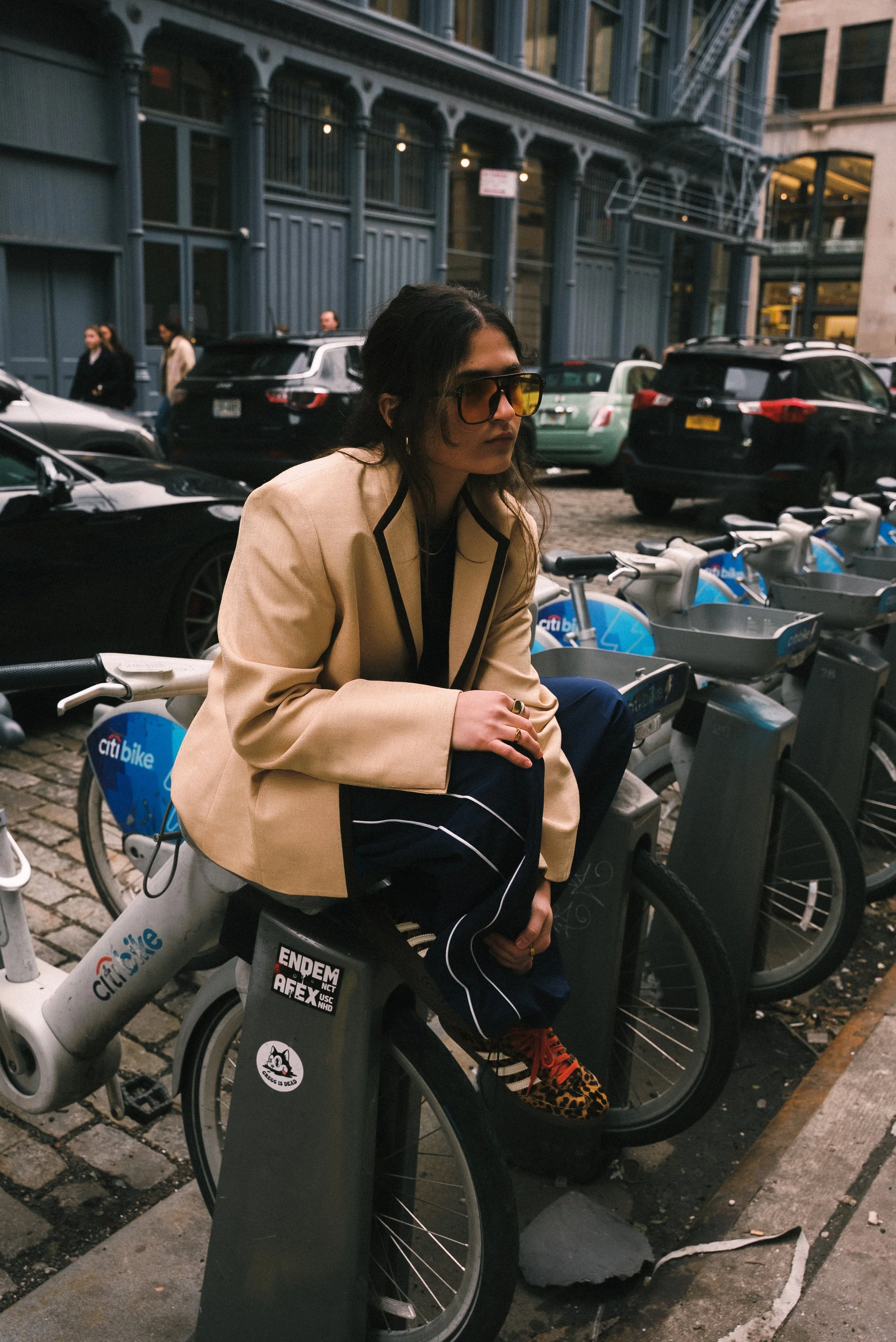 Isa Costa wearing a beige blazer, dark sunglasses, and leopard-print shoes sitting on a Citi Bike in New York City setting with parked cars, bicycles, and a multistory building in the background.