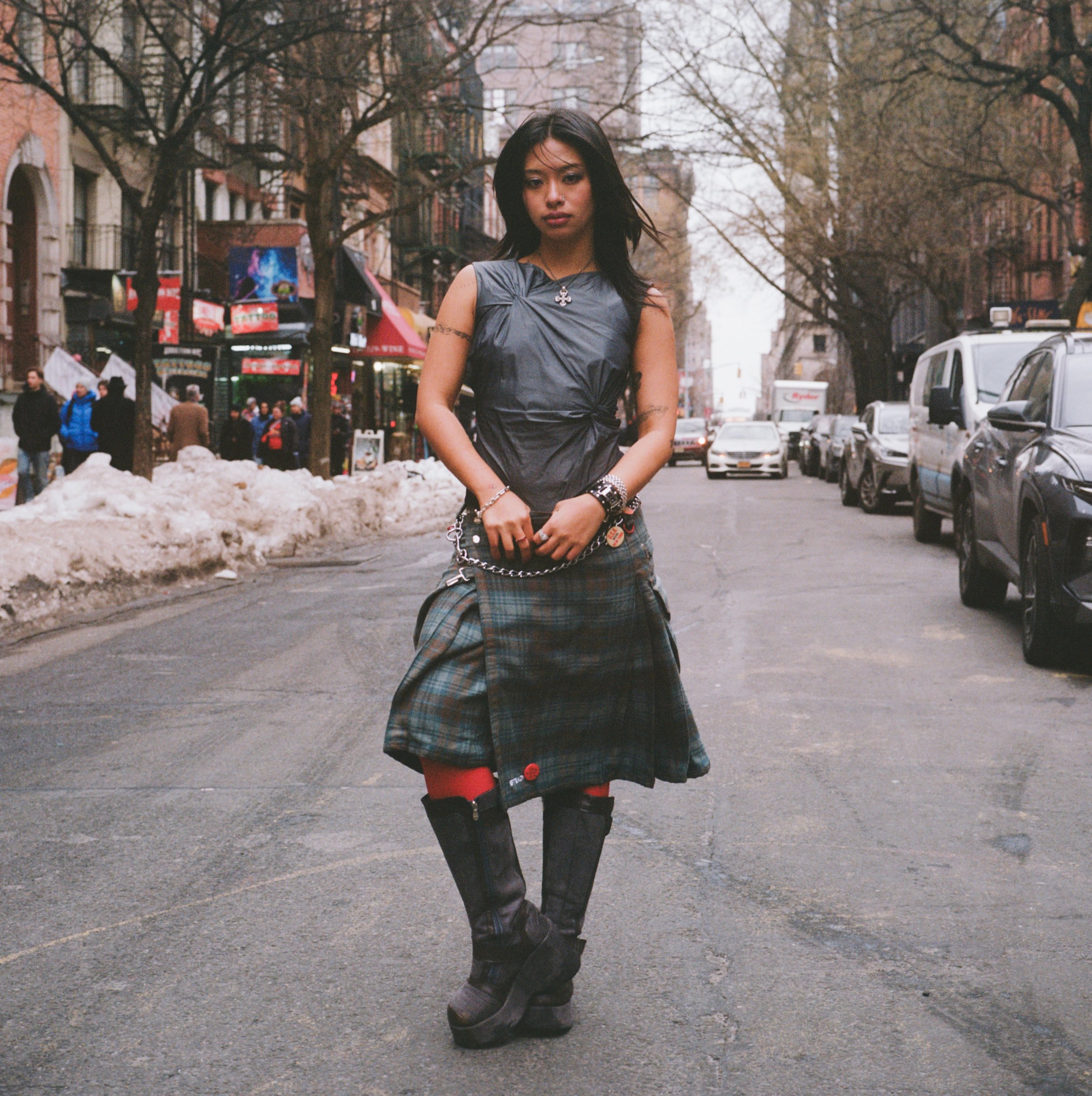 A young woman with black hair styled in a messy look is standing on a city street during winter. She is wearing a black sleeveless top, a plaid skirt, and high-heeled boots. She has accessories including bracelets, rings, and a necklace. The backgrou