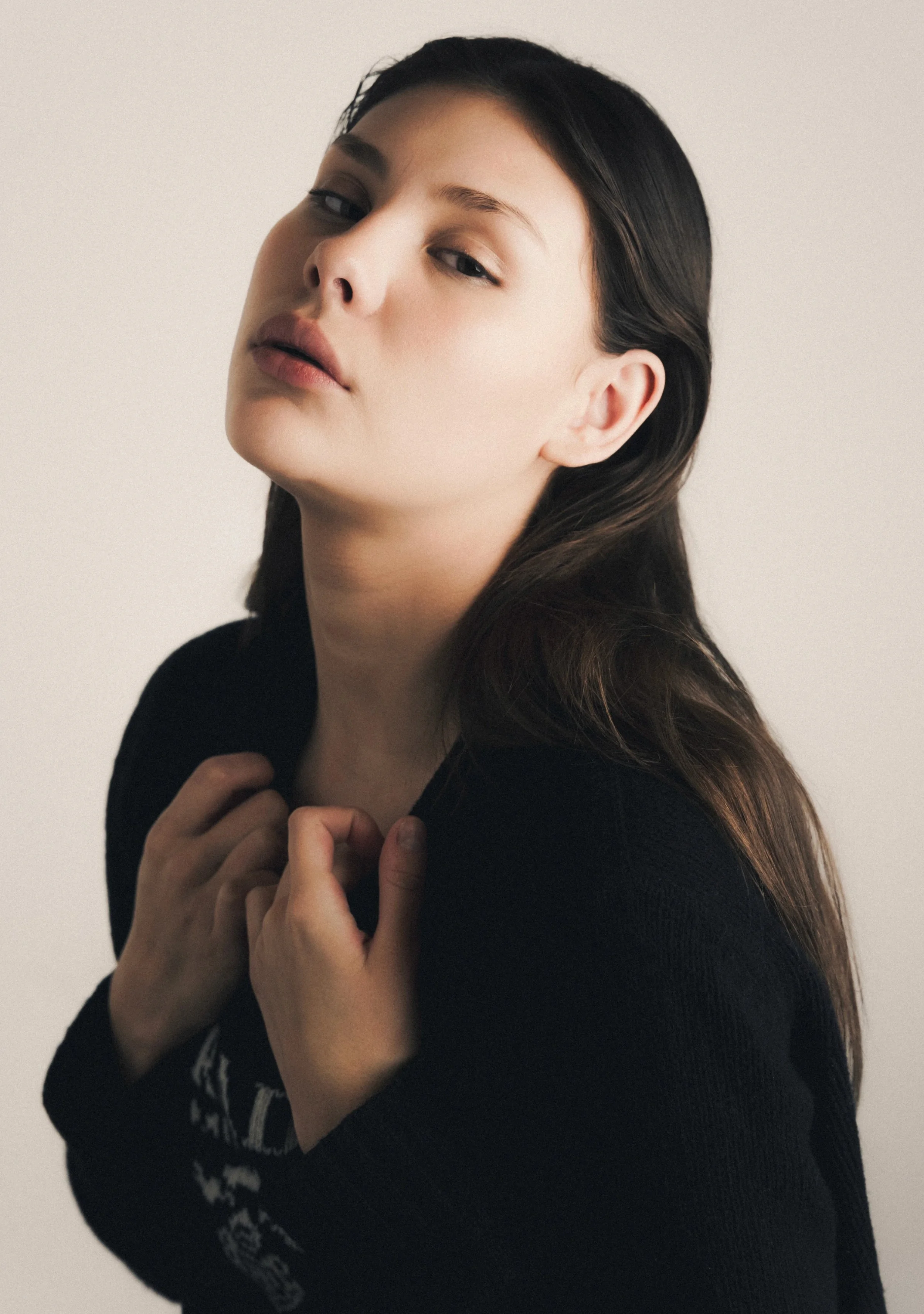 A woman with long, dark hair, wearing a black sweater, looking at the camera with a neutral expression, standing against a plain background.