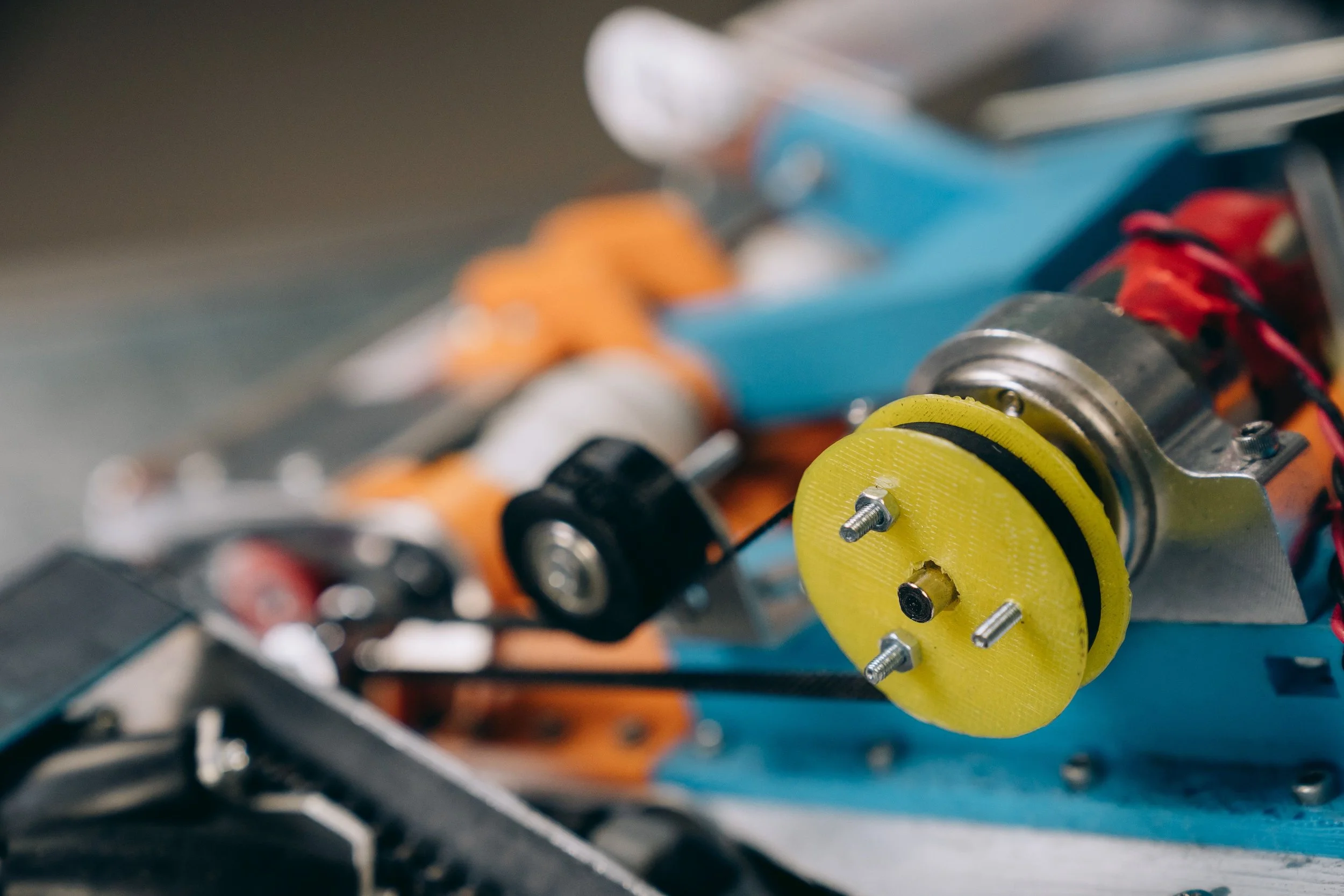 Close-up of a robotic or mechanical assembly with colorful gears and components, including a yellow gear and orange and black parts, mounted on a blue surface.