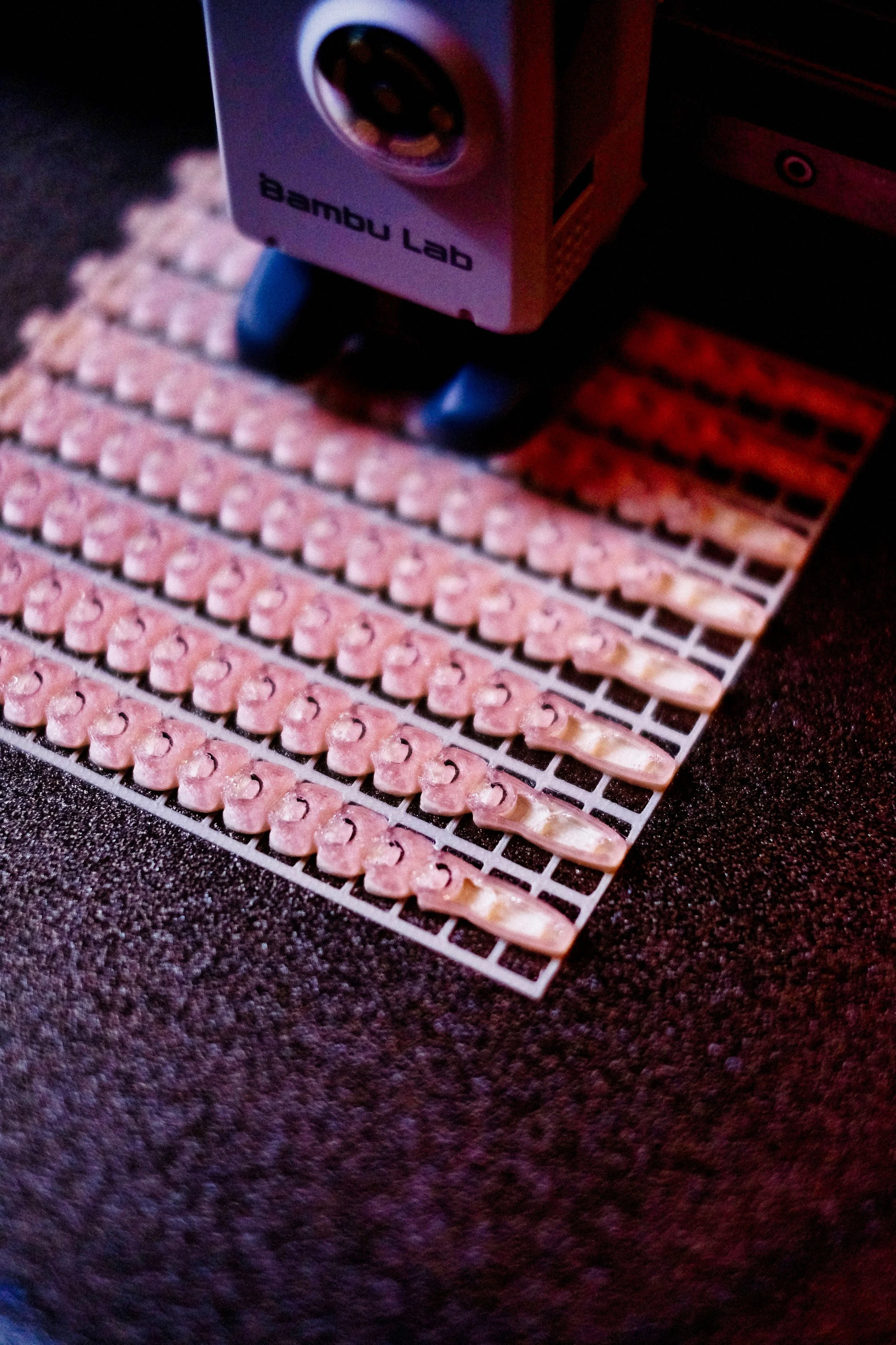 A partial view of a Bambu Lab 3D printer with rows of small white objects on a black surface in front of it.