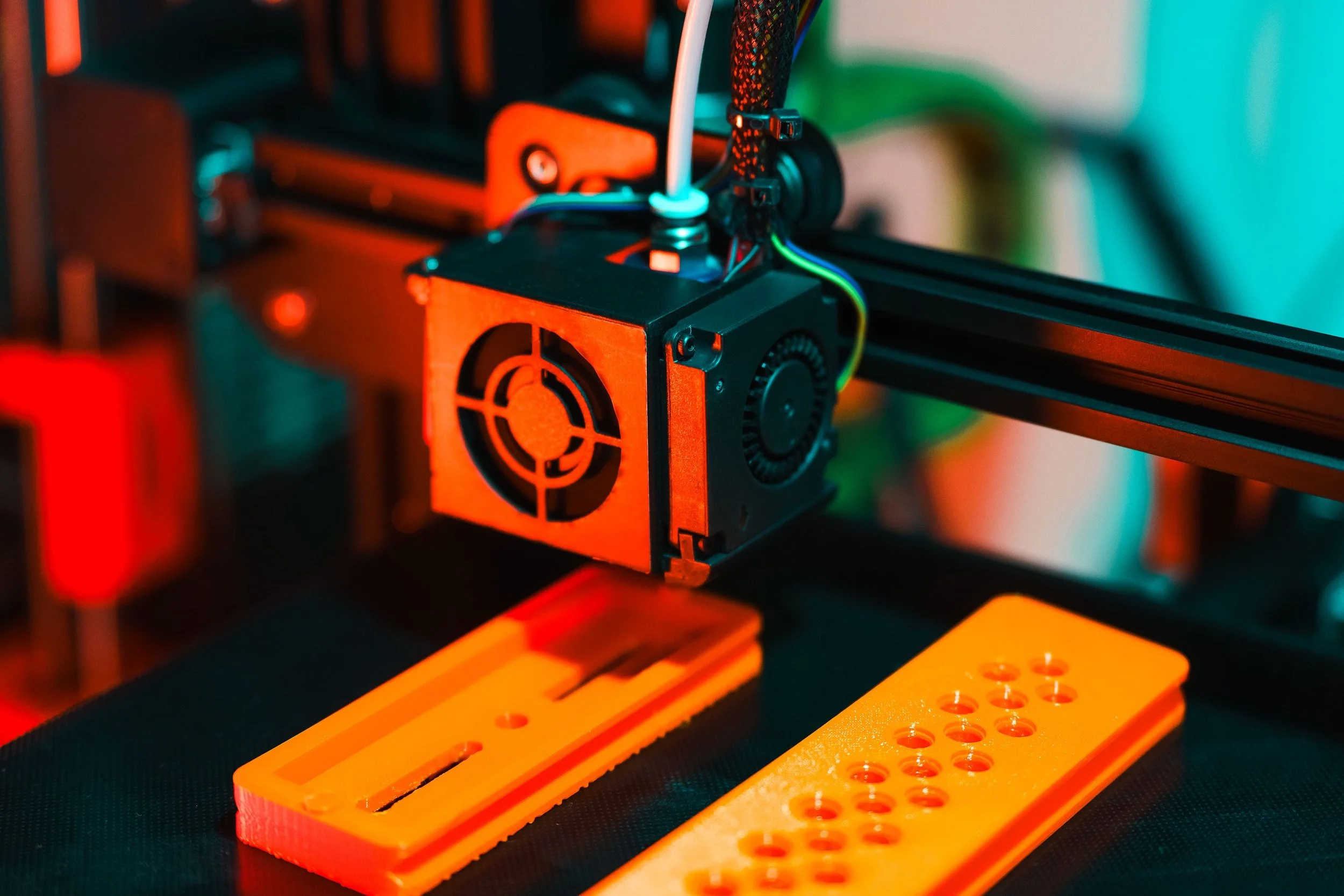 Close-up of a 3D printer's extruder and heated bed, with orange and black components, and colorful wiring, working on a project.