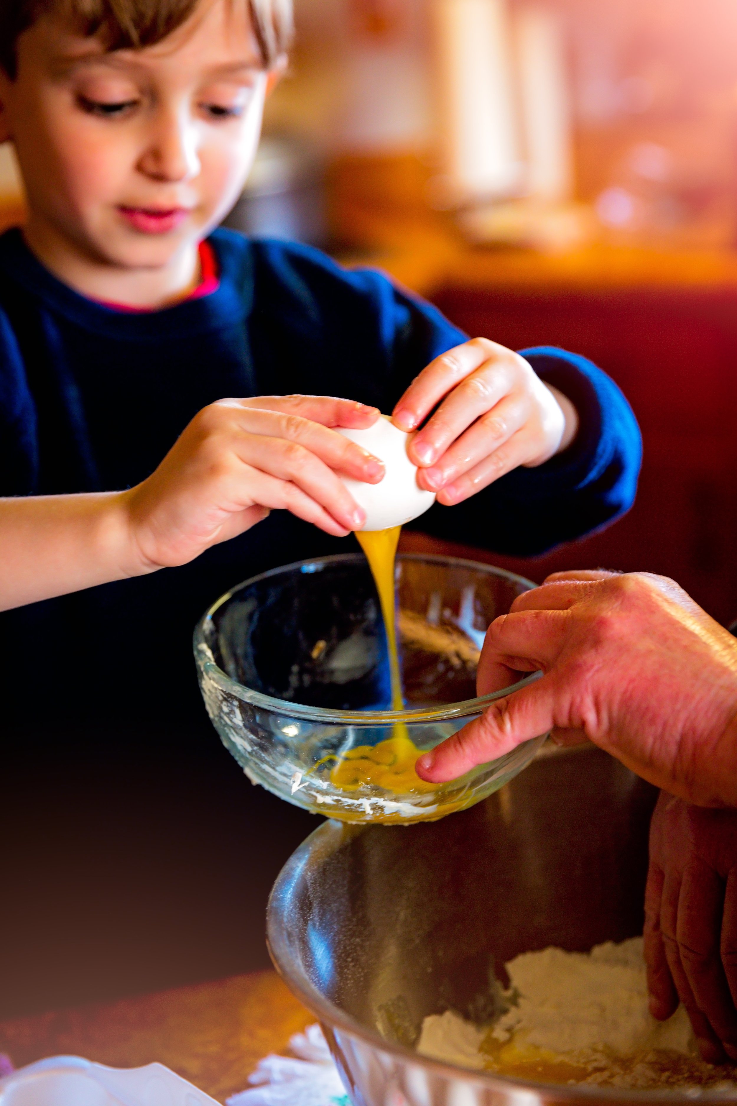 A young boy cracking an egg into a glass mixing bowl while baking. An adult’s hands are holding the bowl. Flour is visible in a larger mixing bowl below.