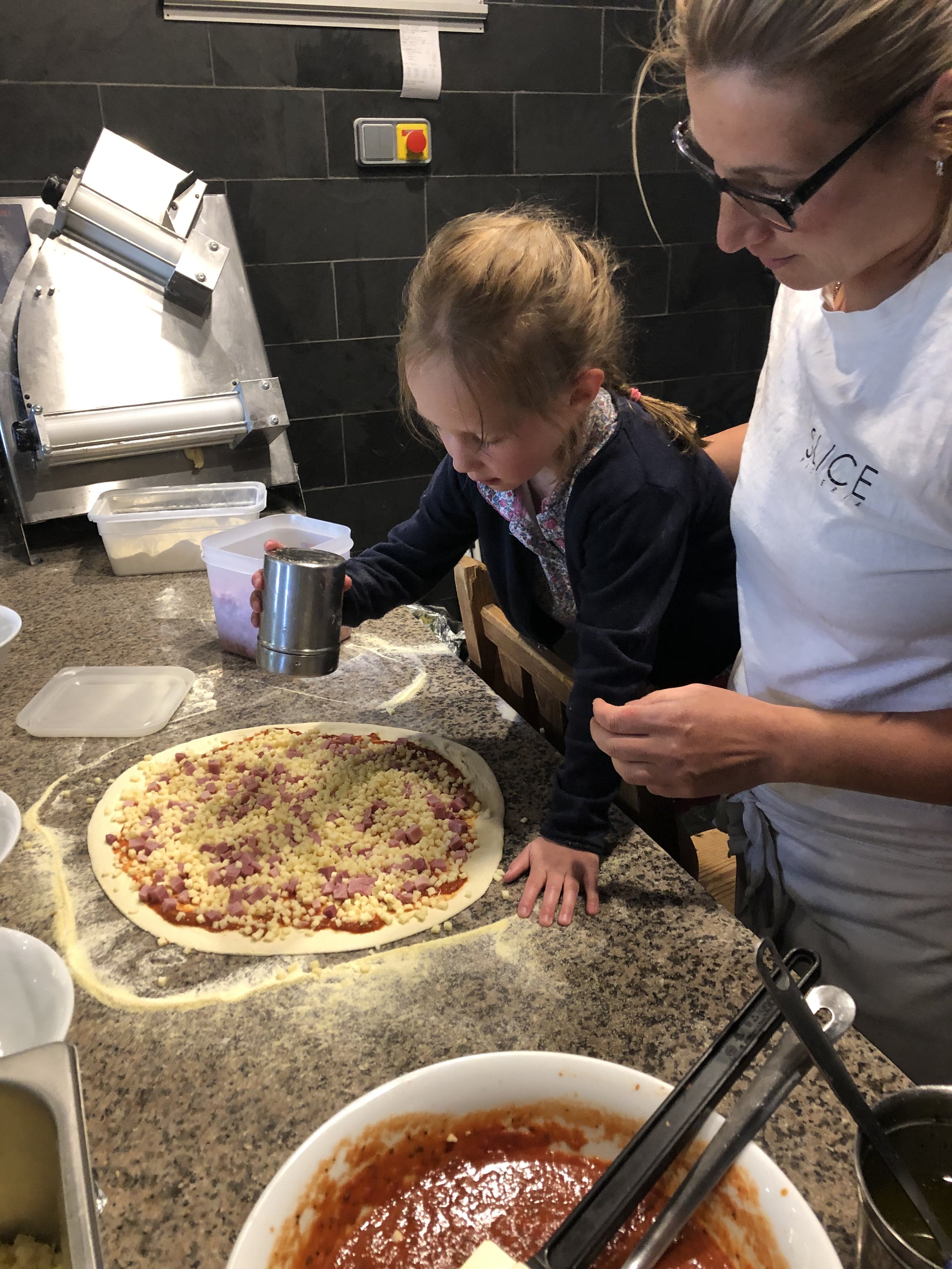 A woman and a young girl preparing to make a pizza, with various pizza toppings and sauce on the table.