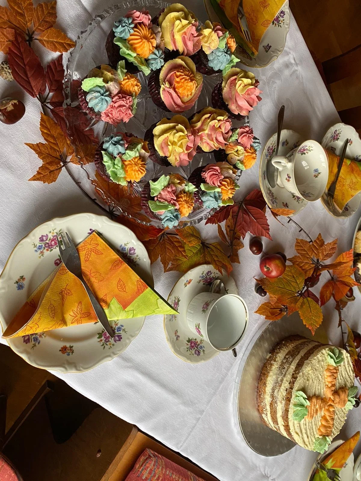 A festive table with cupcakes decorated in colorful swirls, surrounded by autumn leaves, apples, tea cups, and a layered birthday cake with fall-themed decorations.