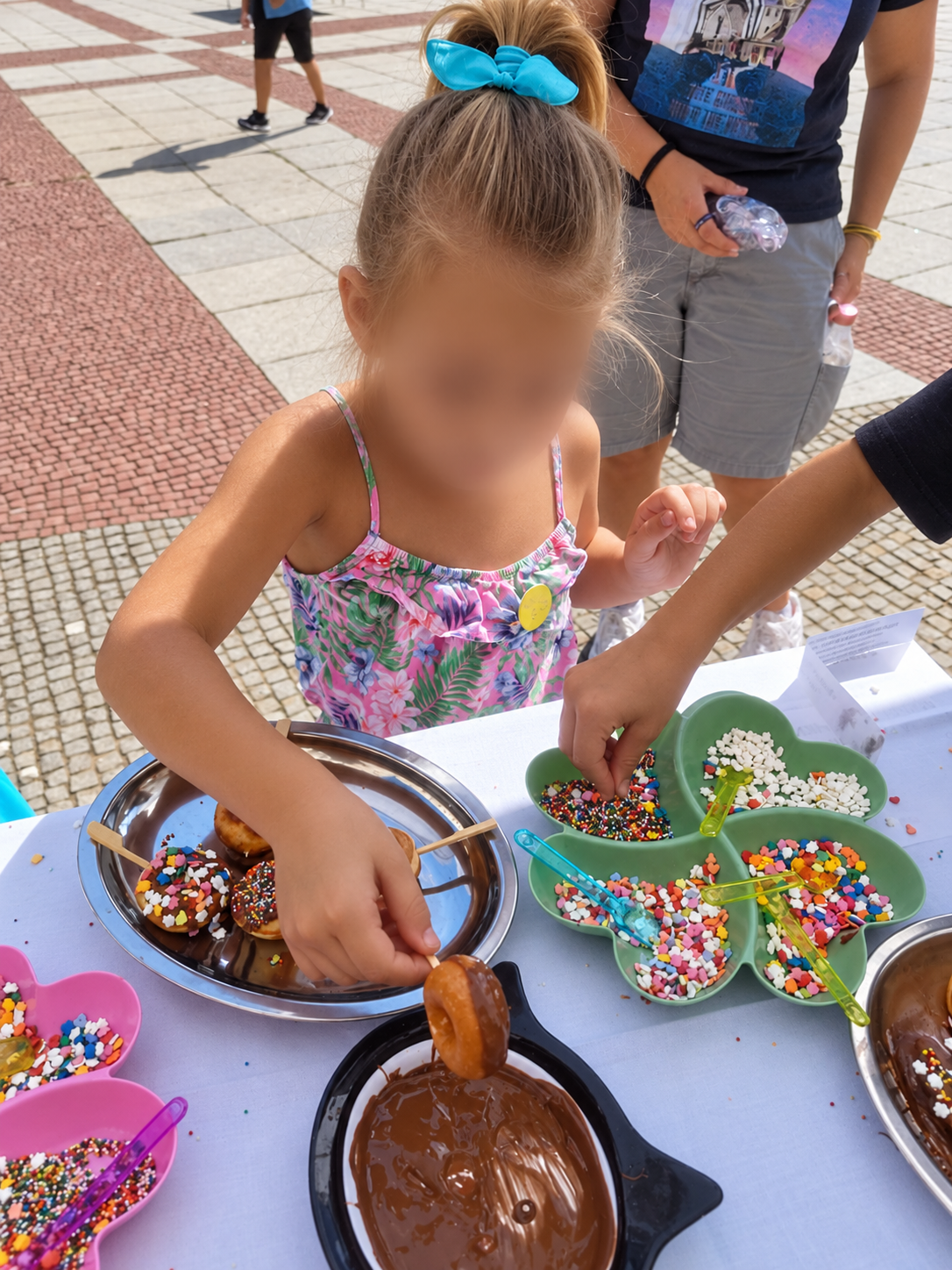 A young girl with a blue bow in her hair dunking a donut into chocolate fondue at an outdoor dessert table filled with colorful sprinkles and toppings.