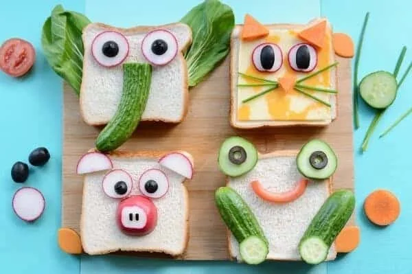 Sandwiches decorated as animals using vegetables and fruits on a wooden board. One looks like a frog with cucumber eyes and mouth, another as a pig with radish eyes and snout, a third as a cat with cheese, carrot ears, and chive whiskers, and the fourth as an elephant with cucumber ears, a radish trunk, and onion eyes.