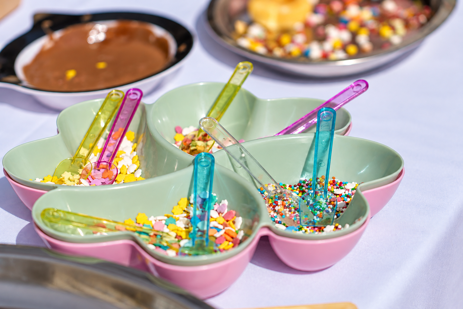 Colorful heart-shaped bowl with various rainbow sprinkles in pink, yellow, white, and multicolored options, with small clear plastic scoops, on a white tablecloth. In the background, there are two other bowls with chocolate and candy toppings.