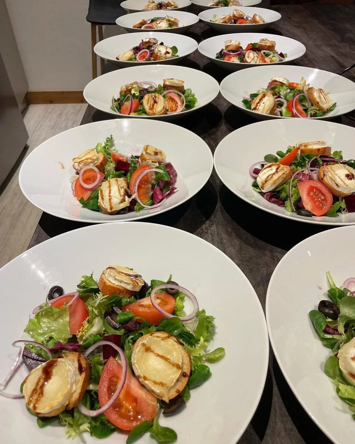 Multiple white plates with salads containing mixed greens, tomato slices, red onion slices, black olives, small grilled cheese sandwiches, and drizzled dressing, arranged on a dark wooden table.