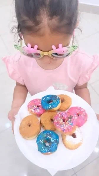 A young girl with reindeer-shaped glasses holding a plate of assorted donuts with colorful icing and sprinkles.